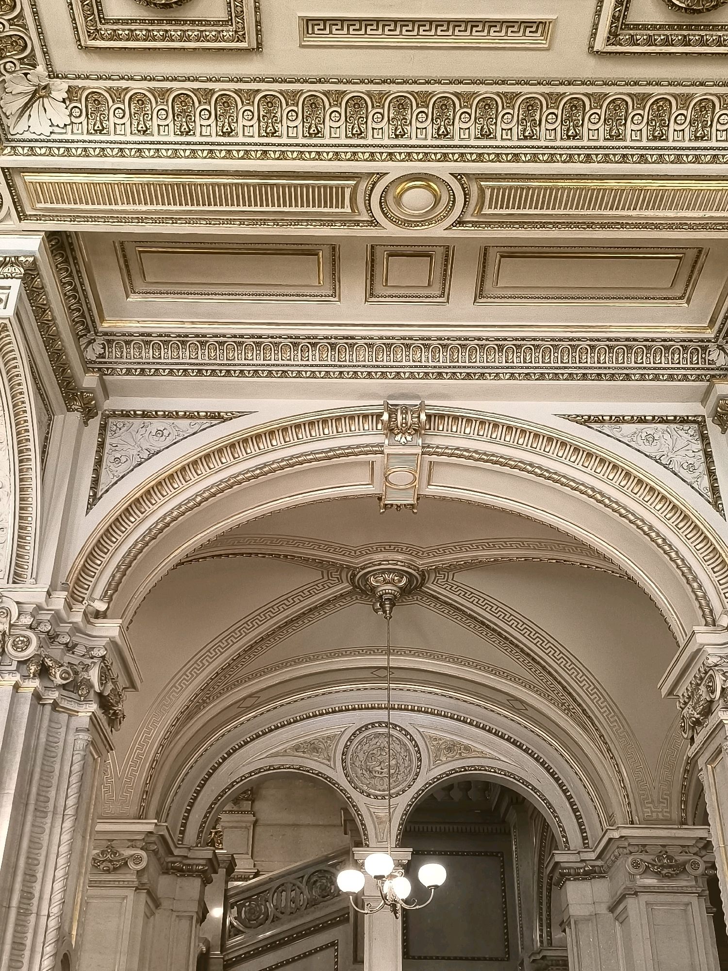 The image shows the opulent interior of the Vienna State Opera. The scene focuses on the ornate architecture of the ceiling and arches, characterized by intricate gold detailing against a cream-colored background. Decorative moldings, friezes with Greek key patterns, and classical motifs are prominently featured. A chandelier hangs from the central arch, casting a soft light on the scene. The architecture suggests a grand and historic building, consistent with the opera house's renowned design. There are no people or visible actions in the frame.