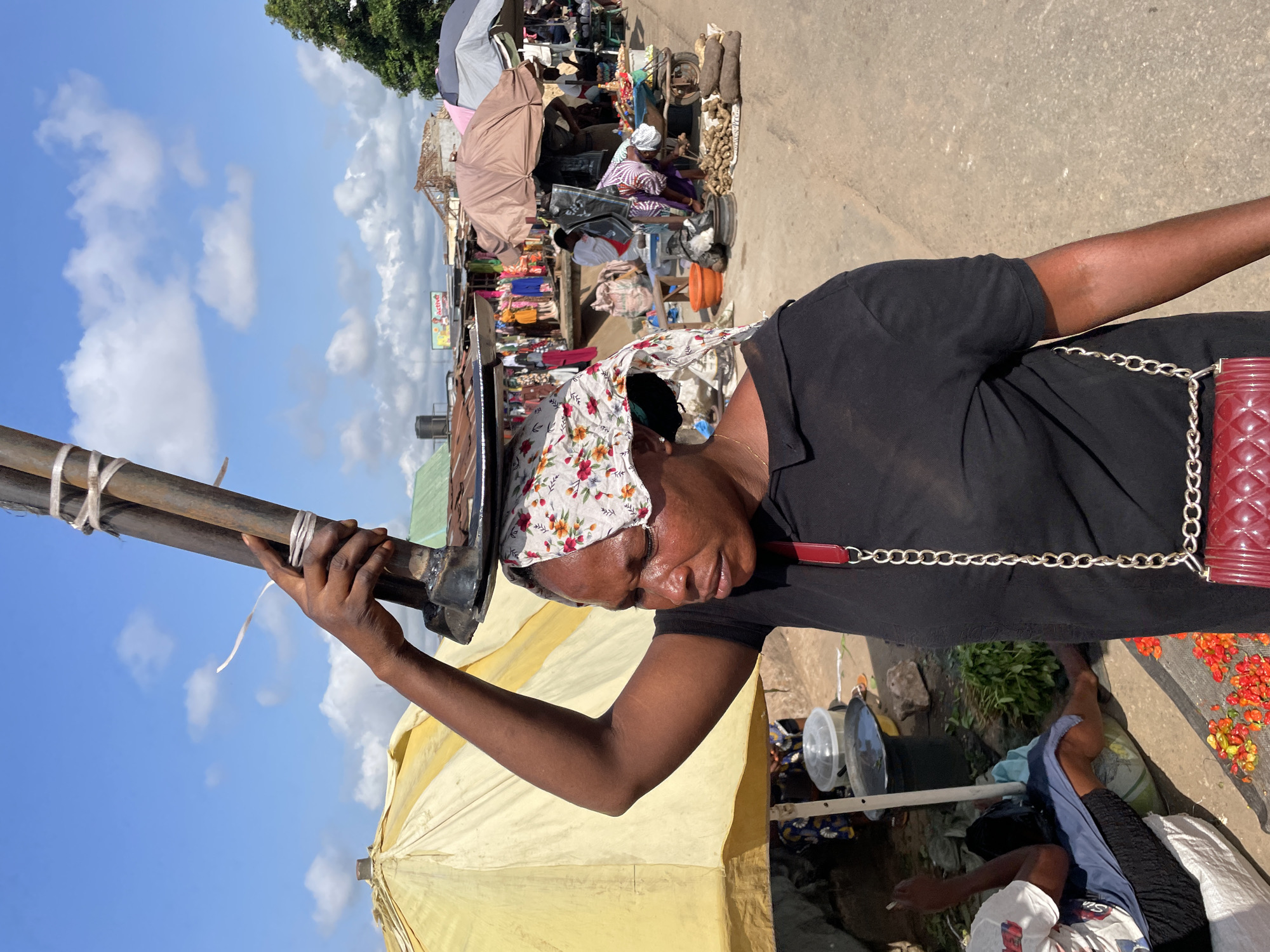 A woman balances a metal object on her head while walking down a dusty street in Onitsha, Nigeria. She wears a black t-shirt and a floral headscarf, with a red purse slung across her shoulder. The street is lined with market stalls, some covered by colorful umbrellas, selling clothing, produce, and other goods. People are seen browsing and tending to their stalls in the background. The sky is blue with scattered white clouds, and trees are visible in the distance. The scene depicts a typical day in a bustling Nigerian market.