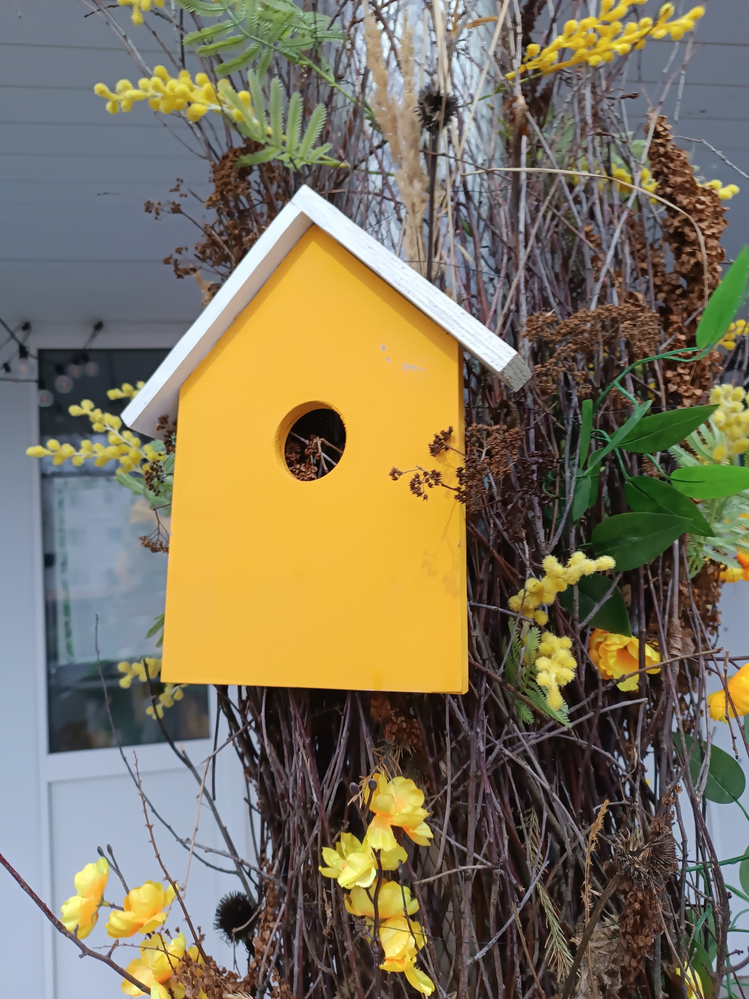 A yellow and white birdhouse with a round entrance hole sits affixed to a bundle of interwoven branches. Yellow flowers and foliage are interspersed among the branches. A section of a light-colored building is visible in the background, with a partially visible window and what appears to be string lights. Based on the prompt, the location is assumed to be Chaikovsky, Russia, though this is not visually verifiable.