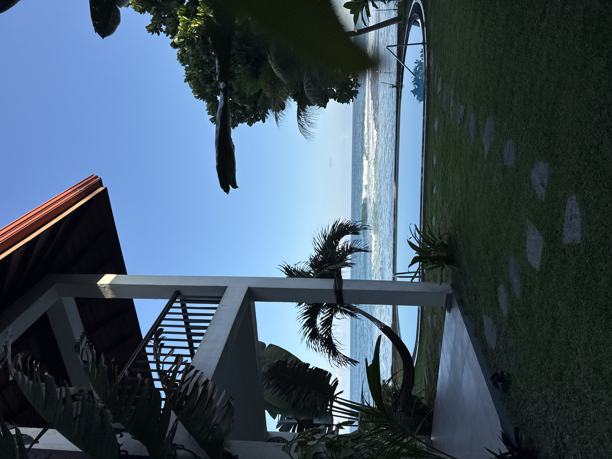 The image depicts an outdoor scene in Goviyapana, Sri Lanka, featuring a multi-story building with a balcony and a thatched roof, adjacent to a lush green lawn. A swimming pool with a silver railing is visible in the foreground, and a hammock hangs between two palm trees. Beyond the lawn and pool, the Indian Ocean stretches to the horizon, with gentle waves breaking on the shore. The sky is a clear, bright blue, indicating fair weather. Tropical foliage, including large palm fronds and dense green trees, frames the scene, suggesting a tropical or subtropical climate. The overall impression is one of a serene tropical resort or private residence by the sea.