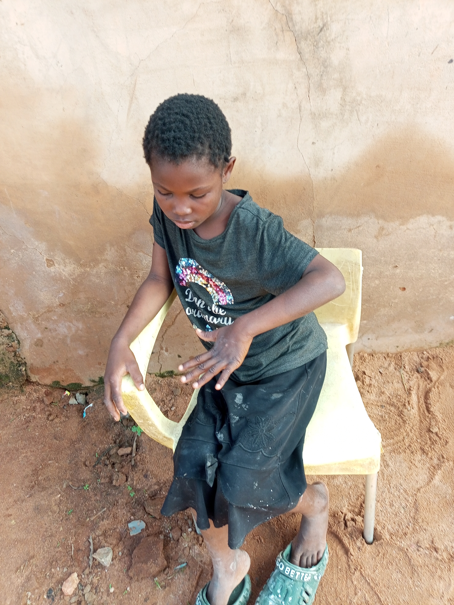 An African girl with dark skin and black curly hair sits on a yellow plastic chair outdoors, facing left with her head bowed and eyes downcast. She is wearing a dark gray t-shirt with a colorful, sparkly design and the words "Don't be ordinary" printed below it in white, and a black skirt. Her hands are resting on the chair. Her bare feet are visible, with one foot wearing a green sandal. The ground is covered in reddish-brown dirt with scattered small rocks and some green weeds. Behind her is a textured, light-colored wall with visible cracks and stains. The scene appears to be in Gombe, Nigeria, likely in an informal outdoor setting.