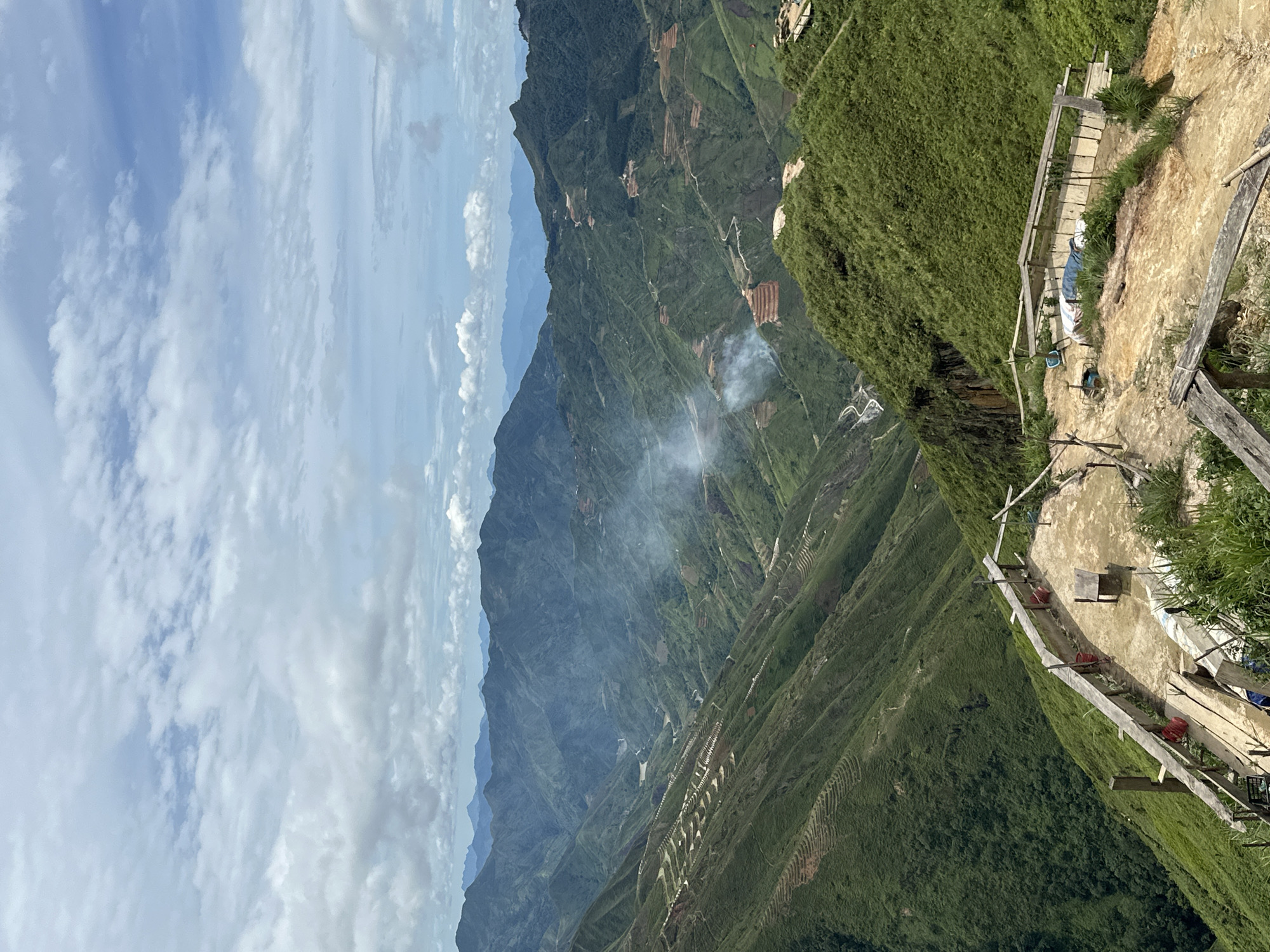 The image captures a panoramic view of a mountainous landscape in Ta Xua, Vietnam, under a cloudy sky. In the foreground, a rustic, elevated wooden platform with a concrete floor overlooks the sprawling green valleys. The platform is equipped with wooden railings and appears to be a viewpoint or resting area, with a small wooden table and seating. Various objects are scattered on the platform, including red buckets and what looks like tarps or bedding.
The middle ground reveals steep, terraced hillsides covered in lush green vegetation, indicative of agricultural cultivation or natural growth. Winding roads and pathways are visible traversing the slopes, suggesting human activity and accessibility within the rugged terrain. Wisps of smoke rise from the valleys, hinting at ongoing activities such as agricultural burning or campfires.
The background is dominated by a series of rolling, verdant mountains that recede into the distance, with a hazy, bluish hue indicating atmospheric perspective. The overall scene depicts a remote, natural environment with evidence of human presence adapted to the mountainous topography of Ta Xua.