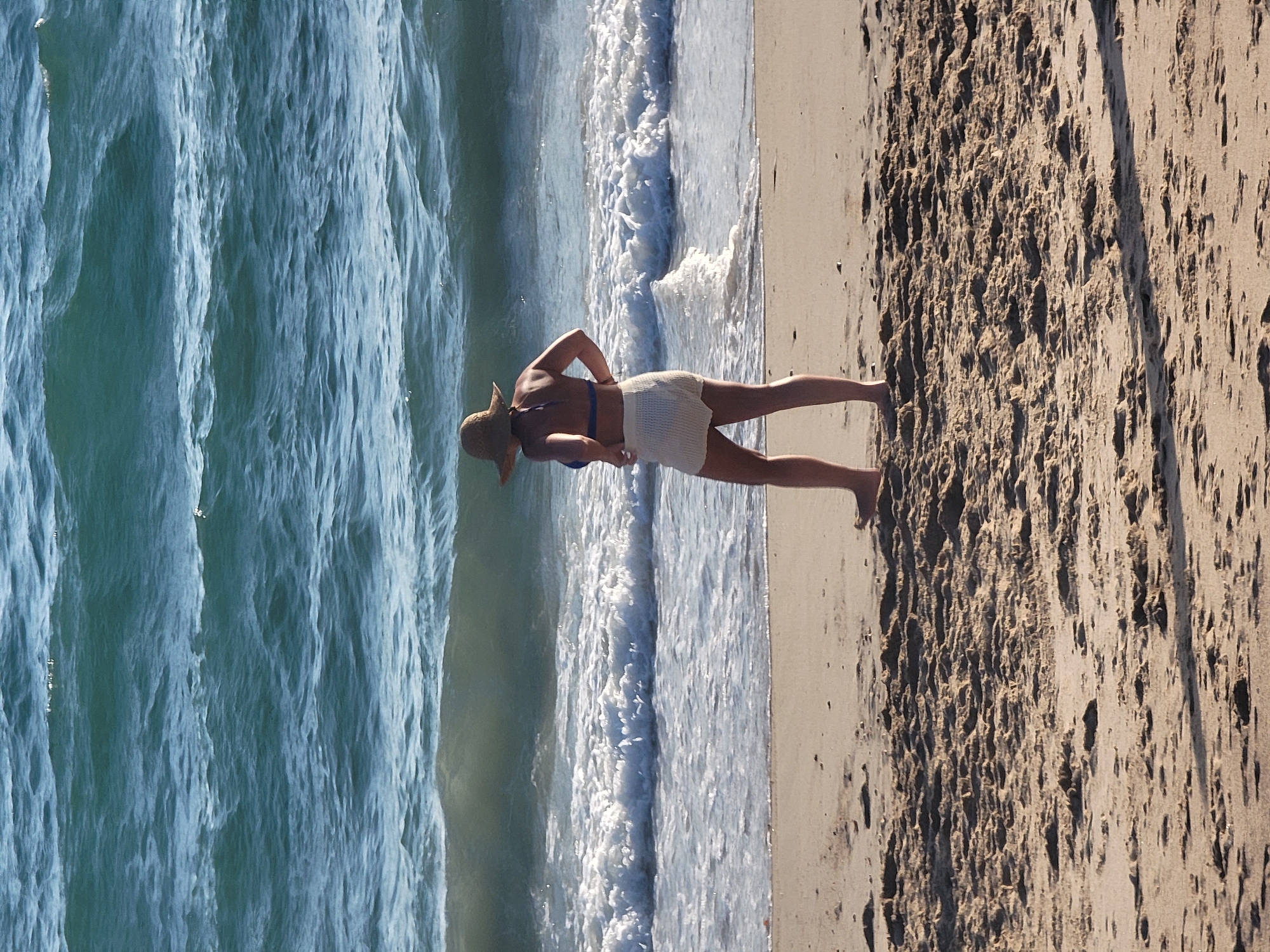 A full-body, vertical shot shows a woman wearing a straw hat, a blue bikini top, and white shorts standing on a sandy beach. She is facing away from the camera, looking at the ocean. The ocean is a deep blue with white-capped waves rolling in. The beach is covered in coarse, light brown sand with some debris and shadows. The image was taken in Quarteira, Portugal.