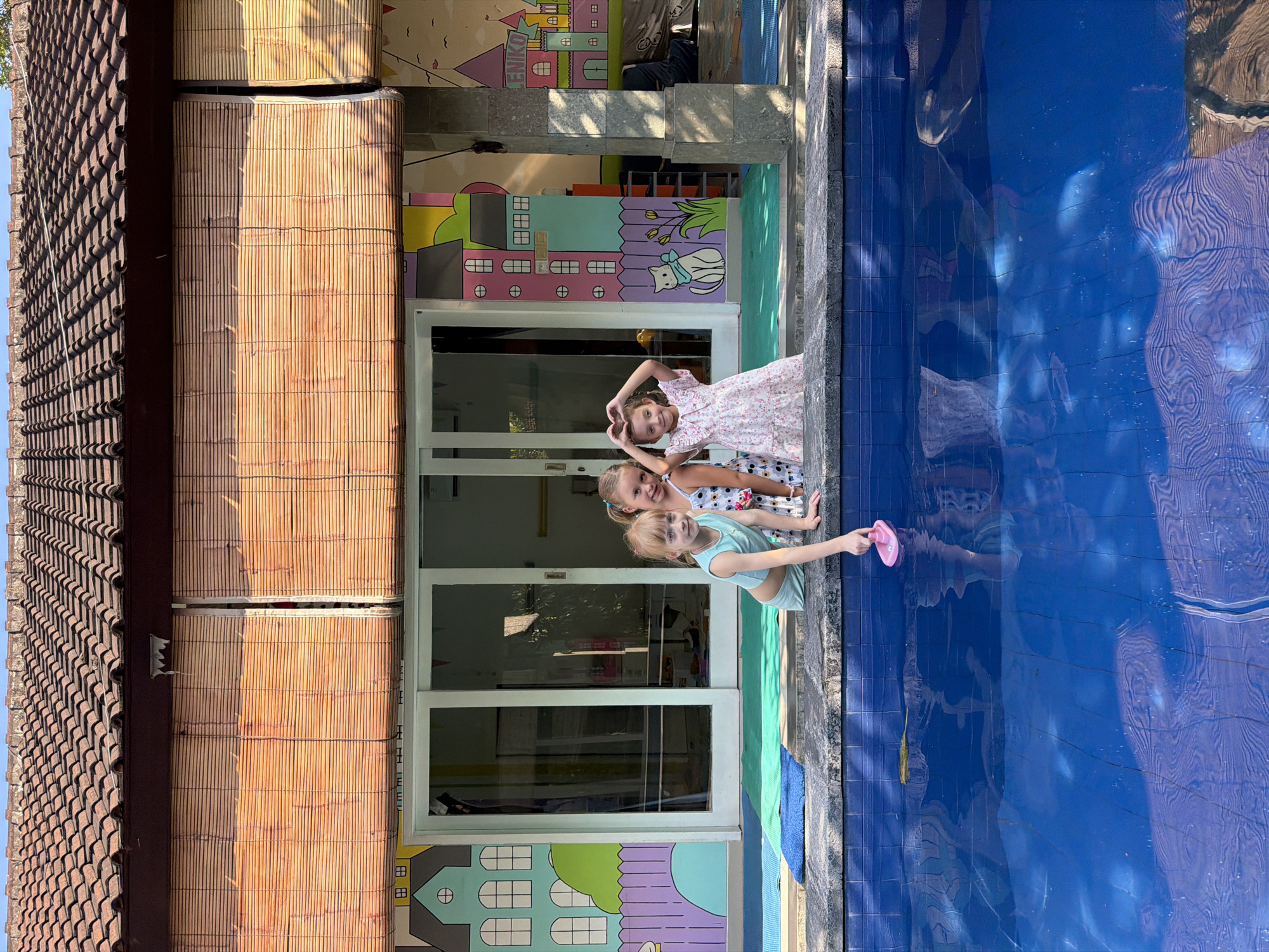Three young girls are standing near a pool. One is reaching into the pool with a pink object. The backdrop is a building with colorful murals of cartoon houses. A set of tan blinds are visible to the left of the girls, and tiled roofing is visible above the blinds. The location is Kuta, Indonesia.