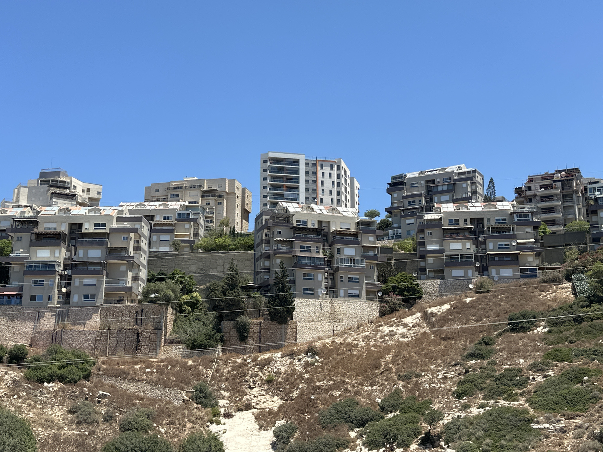 A wide shot captures a residential hillside in Haifa, Israel, under a clear blue sky. The scene is dominated by a cluster of multi-story apartment buildings built into the slope. The buildings are modern, featuring concrete structures with balconies and large windows. Some buildings are clad in light-colored stone or stucco, while others have darker colored sections. Solar panels are visible on the roofs of some of the structures. The hillside itself is characterized by dry, brown vegetation interspersed with patches of green bushes and trees. A series of stone retaining walls are present at different levels of the slope, supporting the built environment. Electrical wires are strung across the foreground, adding a man-made element to the natural landscape. There are no people visible in the image.