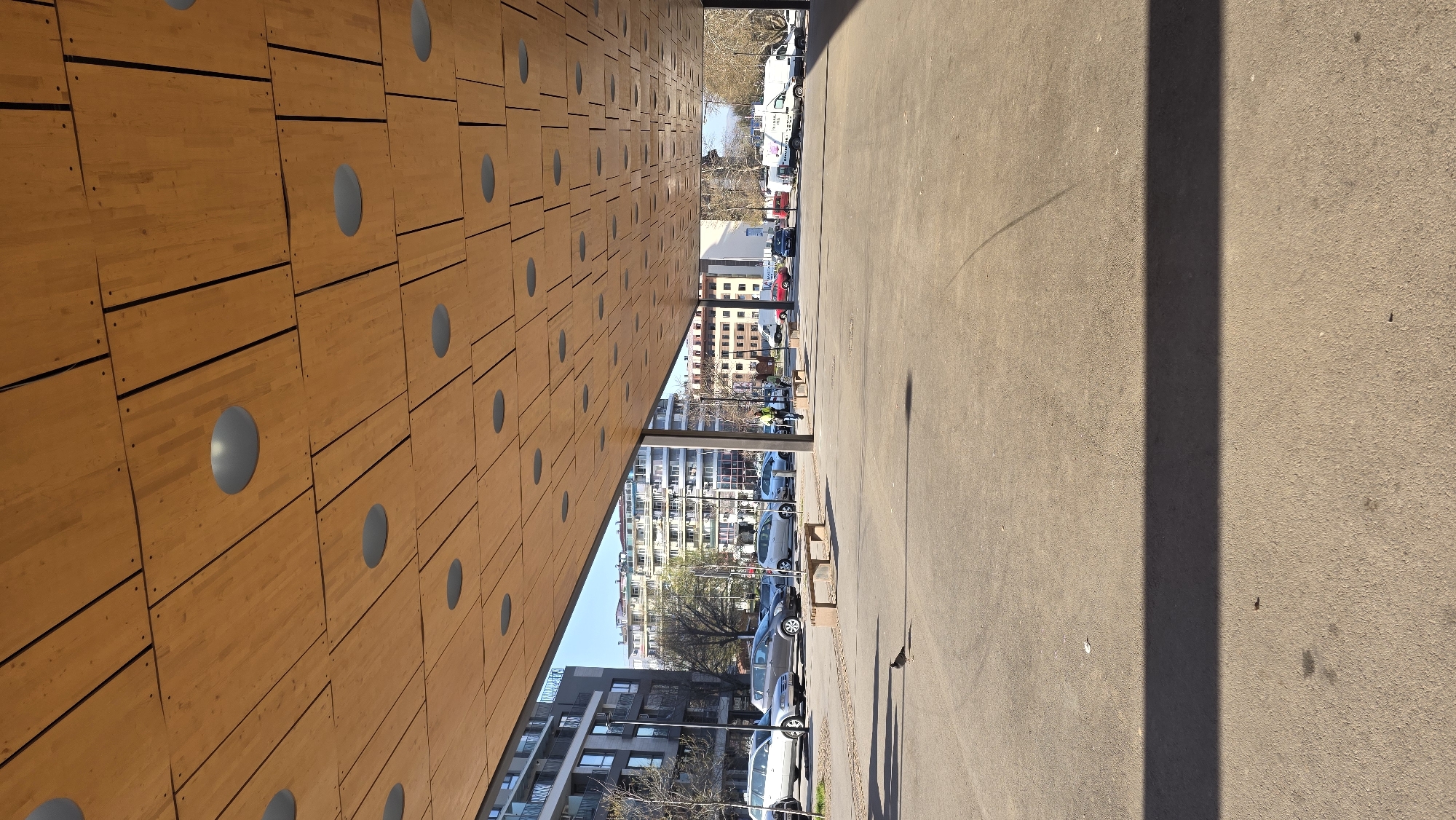 The image depicts an exterior view in Blagoevgrad, Bulgaria, likely along a pedestrian area or walkway. A striking architectural feature dominates the left side, consisting of a wall or overhang constructed of horizontal wooden panels punctuated with evenly spaced, round, recessed light fixtures. This structure creates a covered walkway or sheltered space.
The walkway extends into the distance, revealing a street scene with parked cars, buildings, and people visible in the background. The right side of the image shows the ground, which appears to be paved with concrete or asphalt. A dark, elongated shadow stretches across the surface, possibly cast by a light pole or other vertical object. The overall impression is of a modern architectural space within an urban environment.