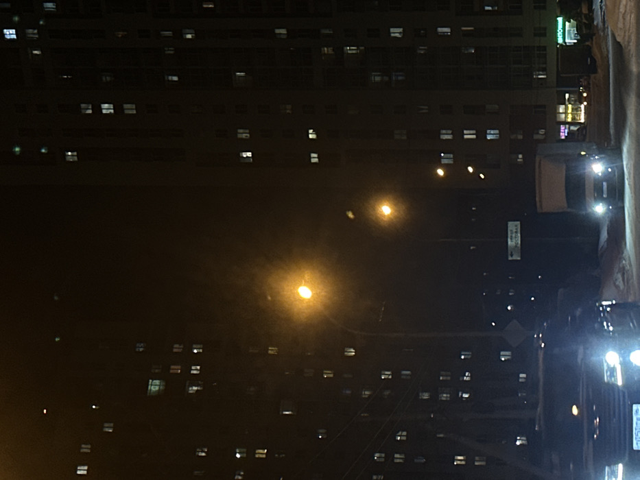 The image shows a dark city street at night. Rows of illuminated windows line tall buildings on either side. Streetlights cast a warm glow. Several vehicles are parked or driving along the road, their headlights adding to the scene's illumination. The overall impression is of a densely populated urban environment after sunset.