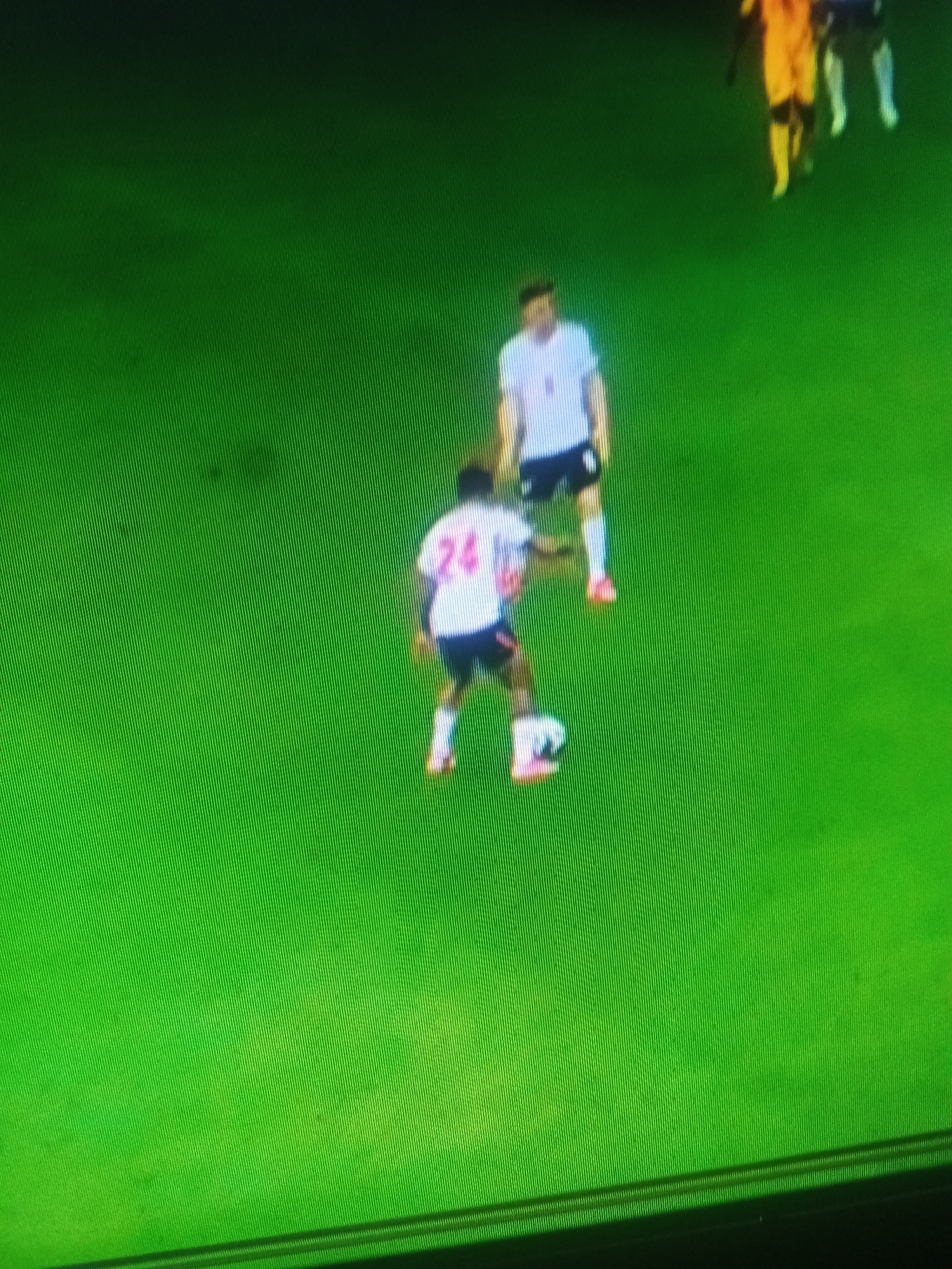 In an aerial, wide shot on a green surface, two male soccer players are visible in the foreground. The player on the left, wearing a white jersey with the number "24" in pink and black shorts, is dribbling a white soccer ball with his right foot. His opponent, standing to his right and slightly behind, wears a white jersey and black shorts, with white socks and pink shoes. Further in the background, two other players are out of focus. The player on the far right wears a yellow jersey and black shorts, while the player next to him is obscured. The green surface appears to be a soccer field. The image is grainy and pixelated, suggesting it was captured on a low-resolution screen. The scene is not identifiable as being in Jalingo, Nigeria.