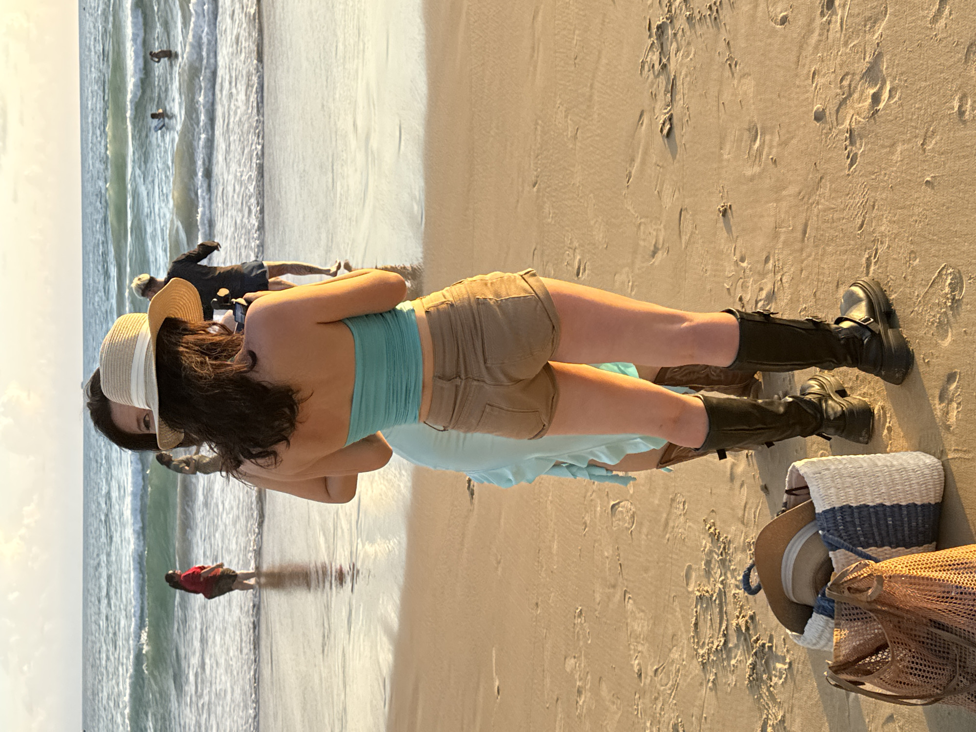 In Karon, Thailand, on a sandy beach with gentle waves, two women stand facing away from the camera, with the ocean and other beachgoers in the background. The woman in the foreground wears a blue strapless top, tan shorts, and black boots. She has a straw hat on her head. Beside her, a second woman is visible, wearing a light blue ruffled dress. In front of them are two beach bags, one woven and tan, the other a striped basket containing a straw hat. In the distance, several people are in the water, with one person on a surfboard. The sky is partly cloudy with warm, golden light, indicating it is either sunrise or sunset.