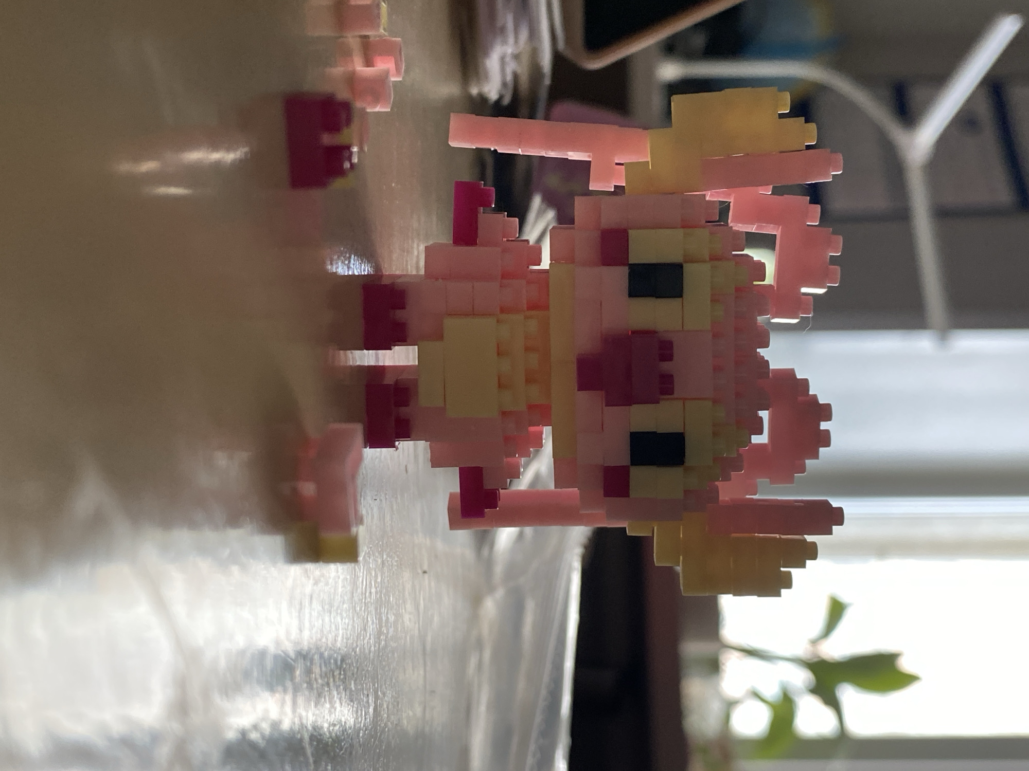 The image, taken with a vertical orientation and appearing rotated 90 degrees counter-clockwise, showcases a small, pixelated figure made from interlocking plastic blocks, similar to NanoBlocks. The figure is predominantly pink and light yellow, with black rectangular blocks forming its eyes and a small red block for what appears to be a mouth or nose. It has a stylized, cartoonish appearance, possibly representing a girl with light yellow hair or pigtails. The body, arms, and legs are constructed from pink blocks, with additional light yellow accents on the torso and feet.
The character stands on a highly reflective, light-colored surface, likely a table or desk, which creates a distorted, blurry reflection of the figure directly beneath it. The background is soft and out of focus. To the right of the figure, a bright, diffused light source, possibly a window, illuminates the scene. A blurred green plant is visible in the lower right portion of the background, and a white, Y-shaped object, potentially part of a lamp or stand, can be faintly seen further back. The lighting suggests the scene is captured during the daytime. The figure is a static object, and no specific activity or event is taking place. There is no visible text in the image. The overall impression is that of a playful, crafted item displayed indoors.