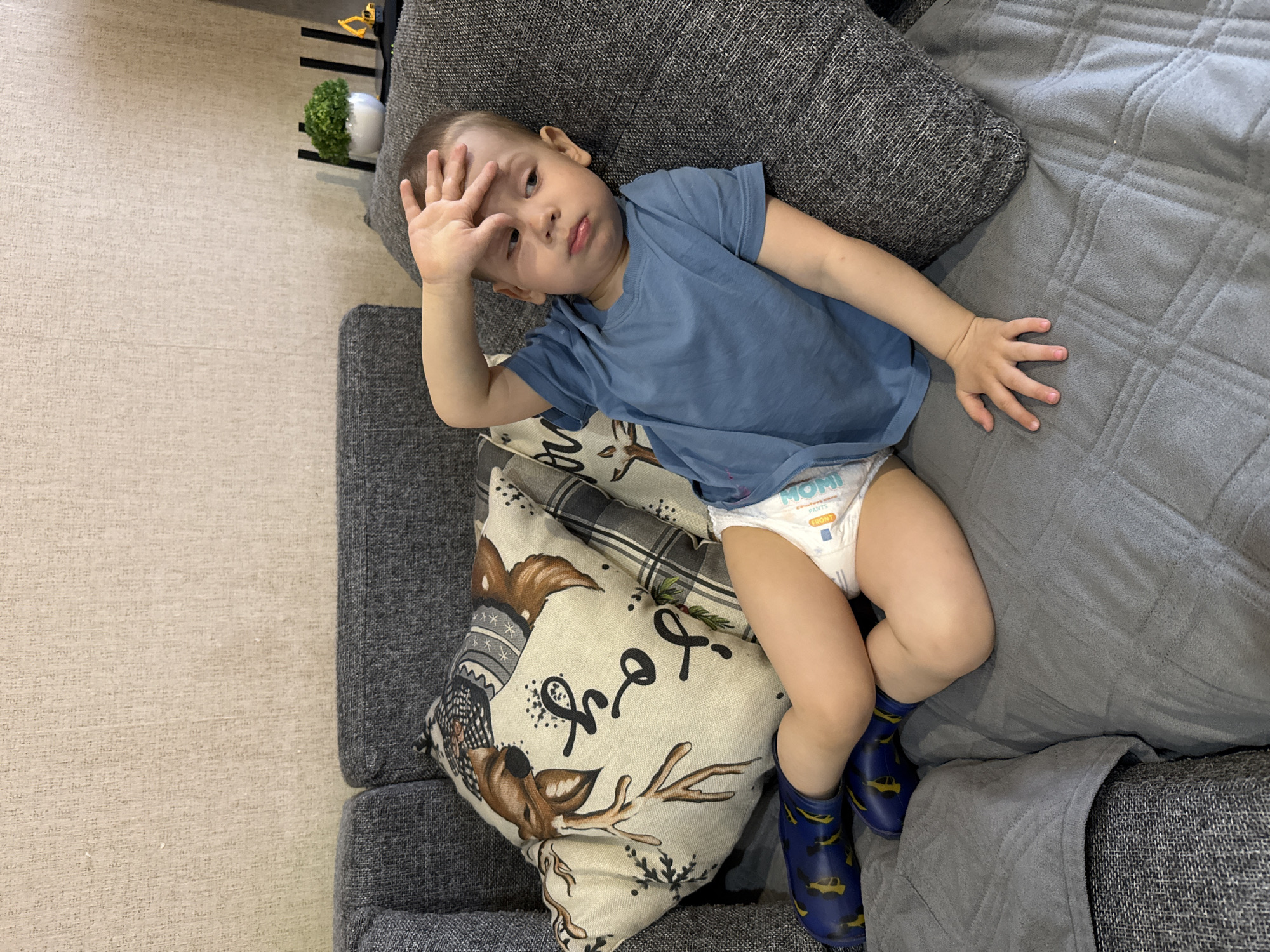 A young child lies on a gray sofa in what appears to be an indoor setting. The child is wearing a blue t-shirt, a diaper, and blue rubber boots adorned with yellow car designs. Their left hand is raised to their forehead. The child is resting on a cushion with a deer design. A small plant and a black object are visible in the upper left corner. The location is Rusanovo, Russia.