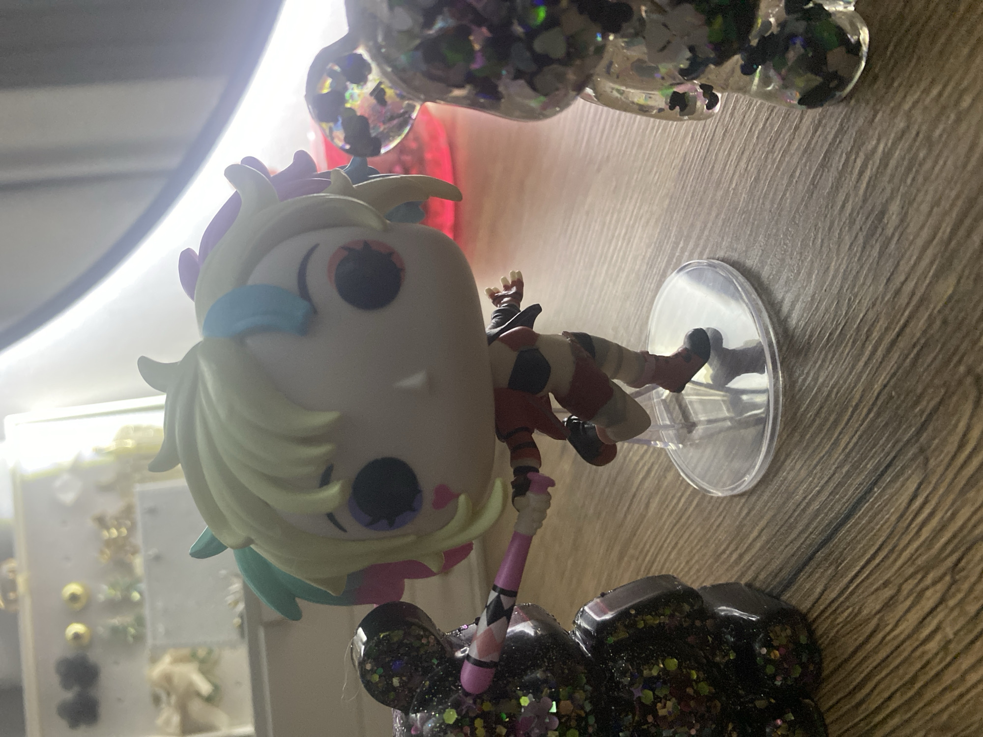 A Funko Pop figure of Harley Quinn stands on a clear plastic circular base on a wooden surface. She has blonde hair with streaks of pink and teal, and is wearing a red and black outfit with red shorts. She is holding a pink and black baseball bat. To her left is a black bear-shaped figurine filled with glitter. To her right is a clear bear-shaped figurine also filled with glitter. In the background, out of focus, are various decorative items on shelves, illuminated by bright lights. The image is taken from a low angle.