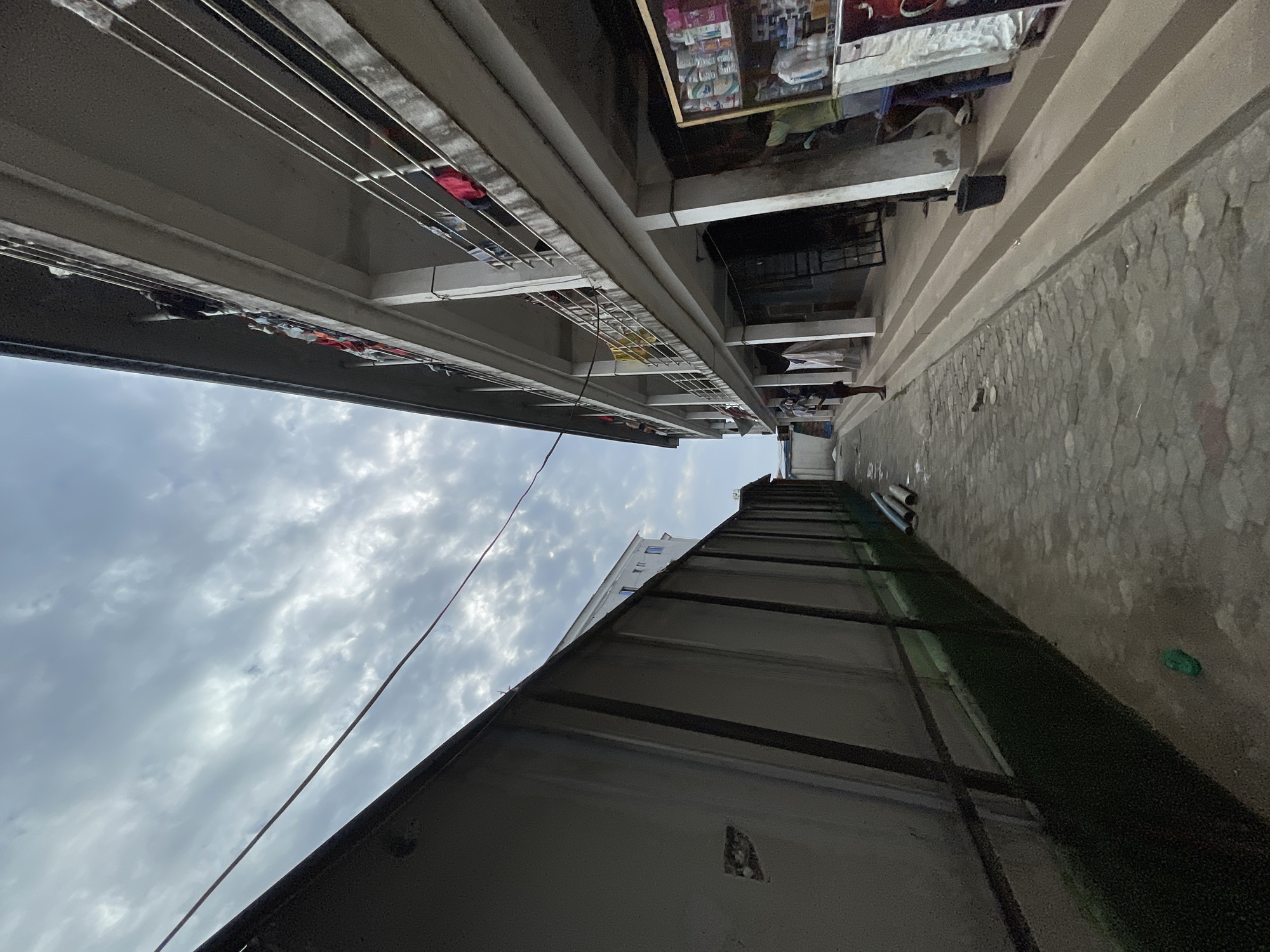 A ground-level, upward-angled photograph shows a narrow alleyway in Ado, Nigeria, flanked by tall buildings on either side. The sky is filled with overcast clouds. Clotheslines with various garments hang between the buildings on the left. The building on the right has a textured stone or concrete wall along the alleyway, and a green, artificial-turf-covered area is visible above it. A thin wire extends from the lower right corner toward the center of the sky. Personal items and supplies are visible in the background. There is a person standing in the background of the alley.
