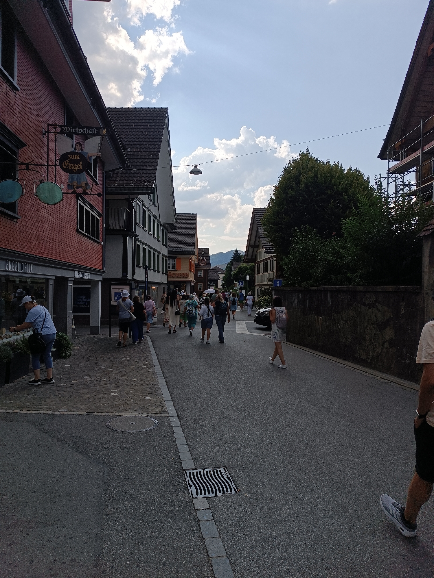 This image depicts a street scene in the town of Appenzell, Switzerland. The foreground shows an asphalt road with a drainage grate on the right side. A person wearing black shorts and light-colored sneakers is walking away from the camera, their right leg extended. The midground features a sidewalk made of cobblestones, with several people walking. To the left of the sidewalk is a brick building with a sign that reads "WIRTSCHAFT Zum Engel" and an illustration of an angel. Further down the street, several other buildings with varying architectural styles are visible, including a building with scaffolding. The background shows trees, hills, and a cloudy sky. Several people are seen walking along the street, some in groups, suggesting a public area or event. The overall atmosphere appears to be casual and active.