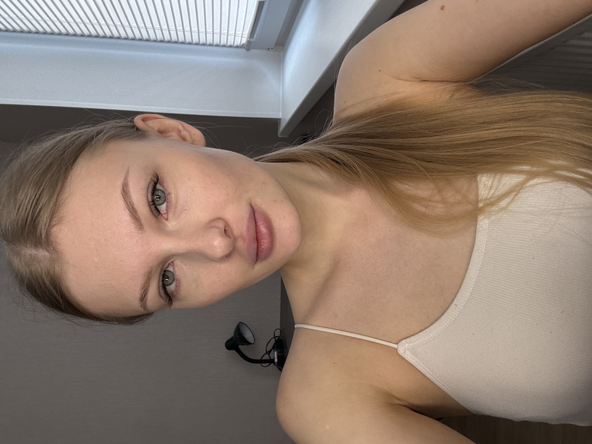 A fair-skinned woman with long, light-brown hair and green eyes poses indoors. She is wearing a white tank top. The background consists of a window with blinds and a wall with a small, adjustable lamp. There is no clear evidence indicating a location within the city of Perm, Russia.