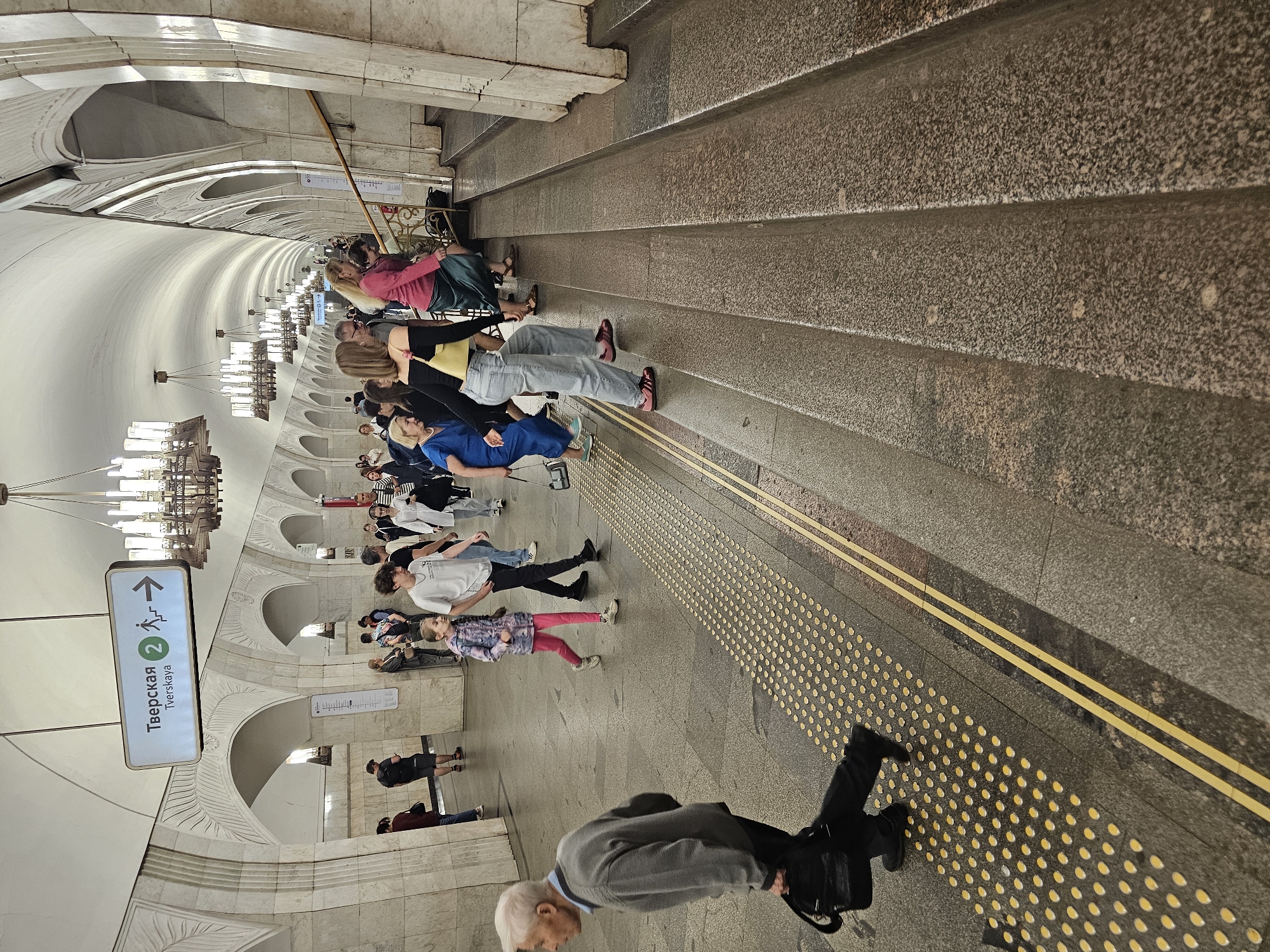 The image depicts a Moscow Metro station, specifically Tverskaya station, as indicated by a sign overhead. The station features grand architecture with arched ceilings and ornate chandeliers. People are seen navigating the space, some walking on the platform and others ascending a set of granite stairs. A young girl in a pink jacket and leggings walks with an older boy in a white t-shirt. An elderly man in a grey sweater carries a black bag as he walks towards the viewer. Several individuals are climbing the stairs, including a woman in a blue dress and a woman in a pink top and long green skirt. The station floor has tactile paving for visually impaired individuals, marked by rows of yellow bumps.