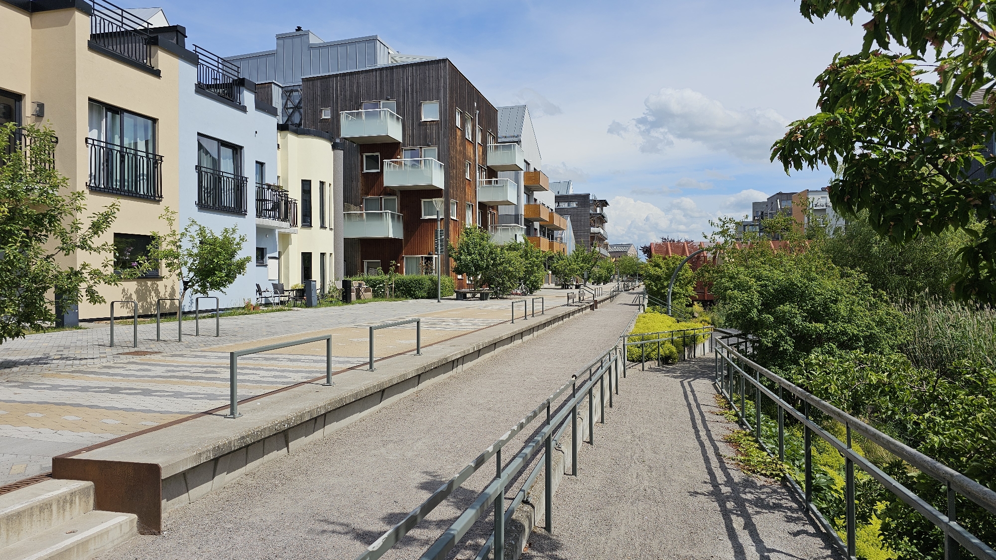 This image depicts the area surrounding the former Åtvidaberg railway line in Linköping, Sweden, which has been redeveloped into a public space. The scene features modern apartment buildings with a mix of light yellow, light blue, and dark wood facades lining a paved walkway. Several multi-story buildings are visible, characterized by balconies with glass railings. A gravel path runs alongside a concrete embankment, separated by metal railings. Trees and lush green vegetation, including bushes and tall grasses, are present on the right side of the path. The sky is blue with scattered white clouds. No people are visible in the image.
