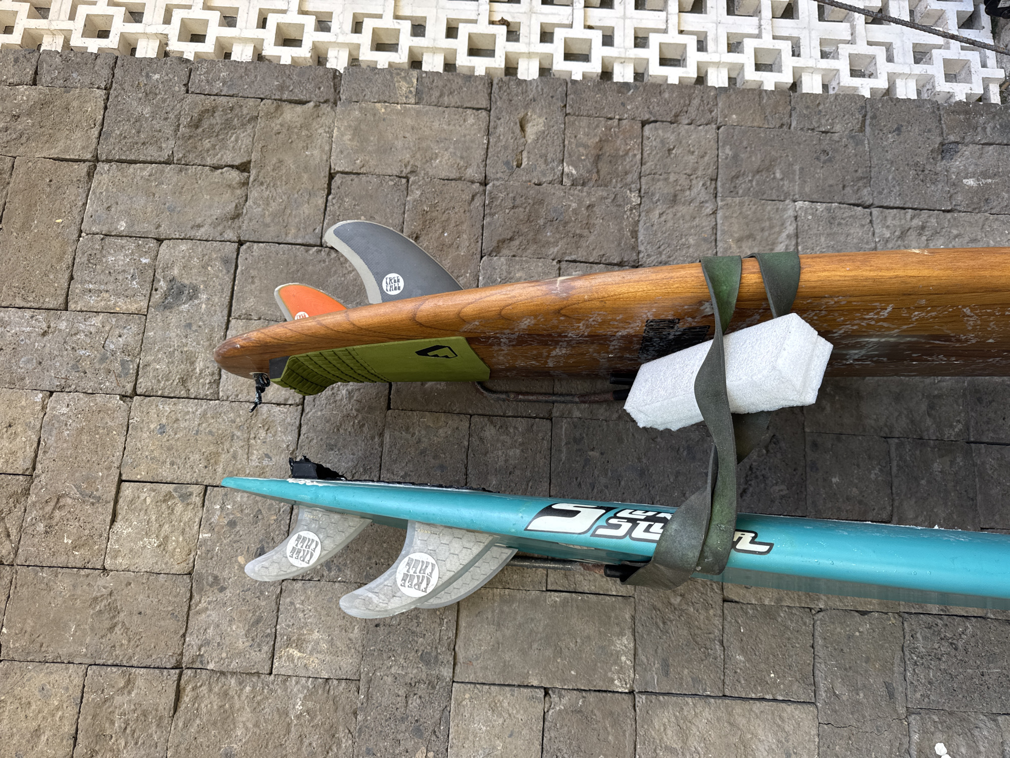 Two surfboards are mounted on a gray brick wall, likely outdoors. The top surfboard is made of wood and has orange and gray fins. The bottom surfboard is teal with white fins. A gray strap secures the boards to the wall. The wall is located in Jimbaran, Indonesia. Above the wall is a white decorative lattice.