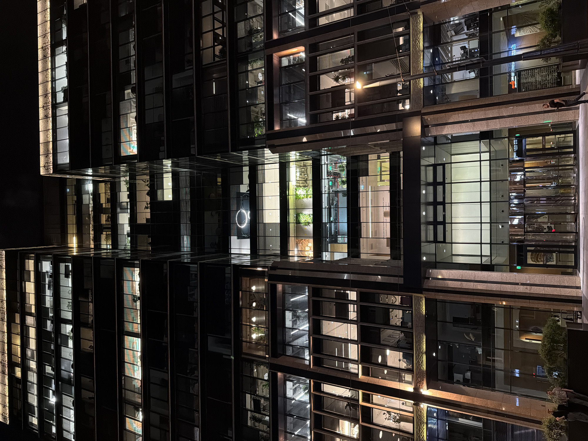The image depicts a modern, multi-story building with numerous windows lit from within at night. The building's facade appears to be a combination of glass and darker, possibly metallic or stone, panels. The architectural style is contemporary, with clean lines and geometric patterns created by the window arrangement.
The image has been rotated, placing the building on its side, which adds to the abstract feel.
Reflections are visible on some surfaces, suggesting wet conditions or reflective materials. There is an outdoor area with vegetation at the base of the building.
The location appears to be in Moscow, Russia.