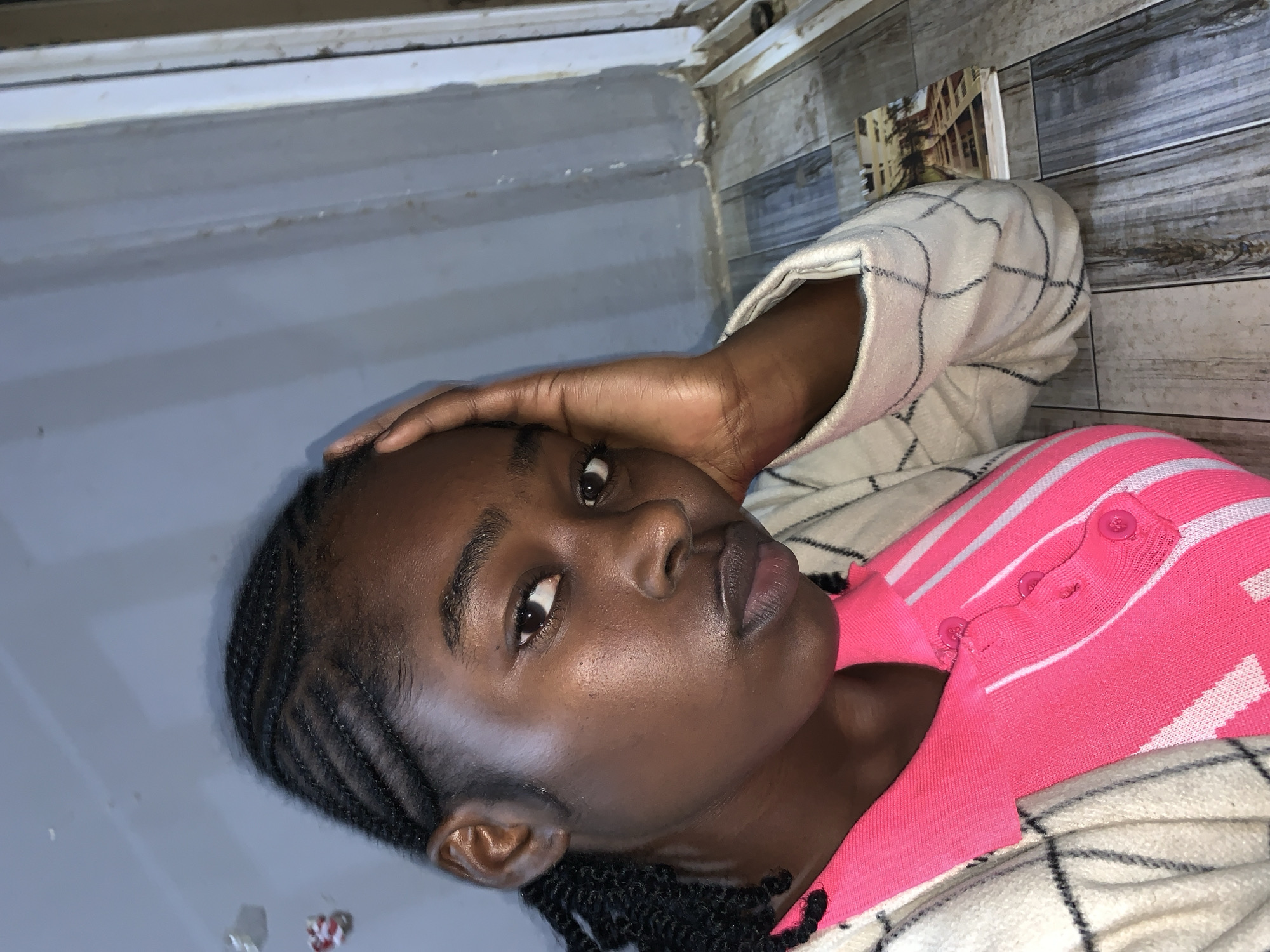 An image taken indoors in New Stadium, Nigeria, depicts a young Black woman in a close-up, angled slightly towards the right. She has her hair styled in cornrows, pulled back from her forehead. She is wearing a bright pink knit polo shirt with white horizontal stripes and three visible buttons down the placket, layered under a cream-colored, possibly quilted, cardigan featuring a dark checkered pattern. Her left hand is raised, with fingers curled and resting against the left side of her face and temple. She looks directly forward, making eye contact with the viewer. The background consists of a light gray paneled wall, with a white vertical frame visible along the right edge, suggesting a doorframe or window casing. The floor beneath her is composed of distressed, light gray and brown wooden planks. A small book or notepad with a landscape image on its cover is visible on the floor to the lower right.