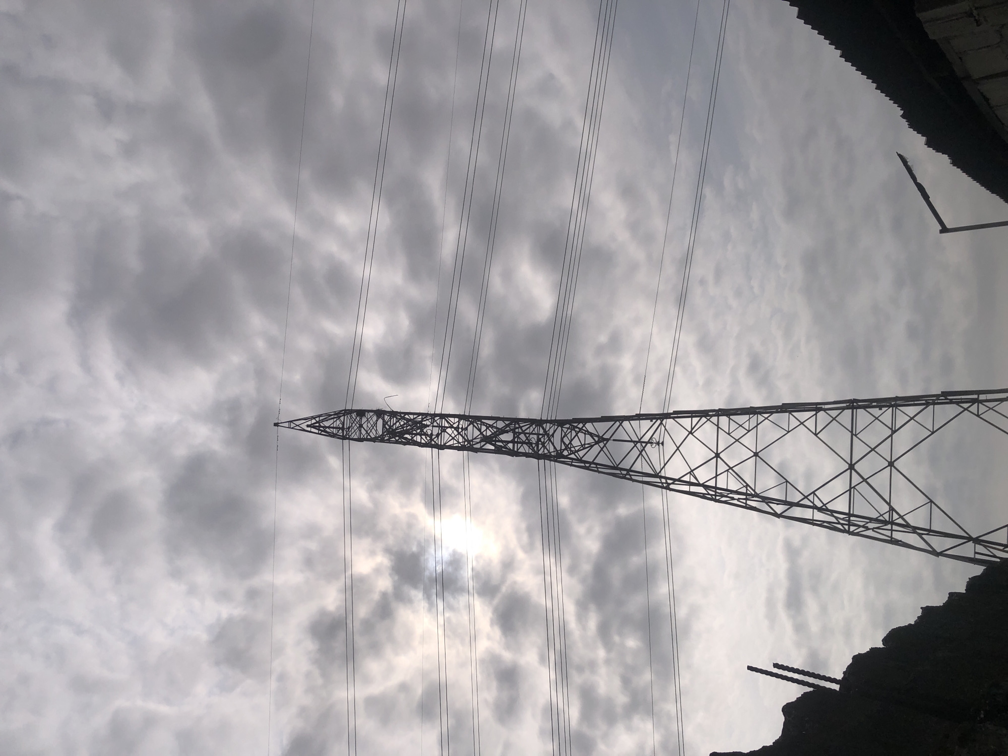 The image depicts an overcast sky with scattered clouds, through which the sun is partially visible, casting a bright glow. A large, metal electricity pylon dominates the center of the frame, its lattice structure reaching upwards. Several power lines are strung across the sky, supported by the pylon. In the foreground, on the left, a dark, jagged structure, possibly a wall or debris pile with rebar protruding, is silhouetted against the sky. On the right side of the image, a corrugated roof structure is visible along with a street light pole. The overall scene appears to be an industrial or semi-urban environment. The location is Ado, Nigeria.