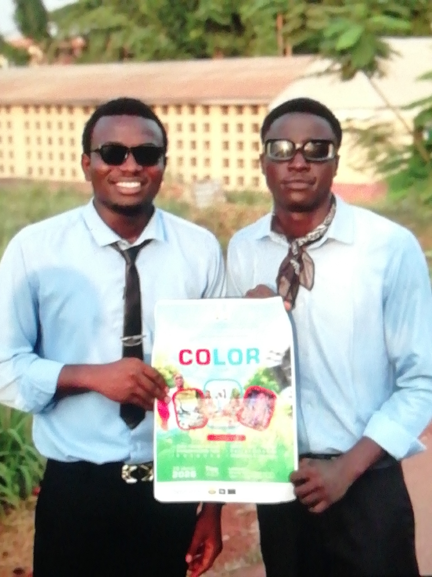 Two men stand side-by-side outdoors, holding a poster titled "COLOR". The man on the left wears sunglasses, a light blue collared shirt, a black tie with a silver clip, and black pants with a silver belt buckle. The man on the right also wears sunglasses and a light blue collared shirt, paired with a patterned neckerchief. Both are facing the camera. The background includes a large building with multiple windows and green vegetation. It is likely this photograph was taken in Enugu, Nigeria, given the request specified that location. The men are likely promoting or advertising the “COLOR” poster.