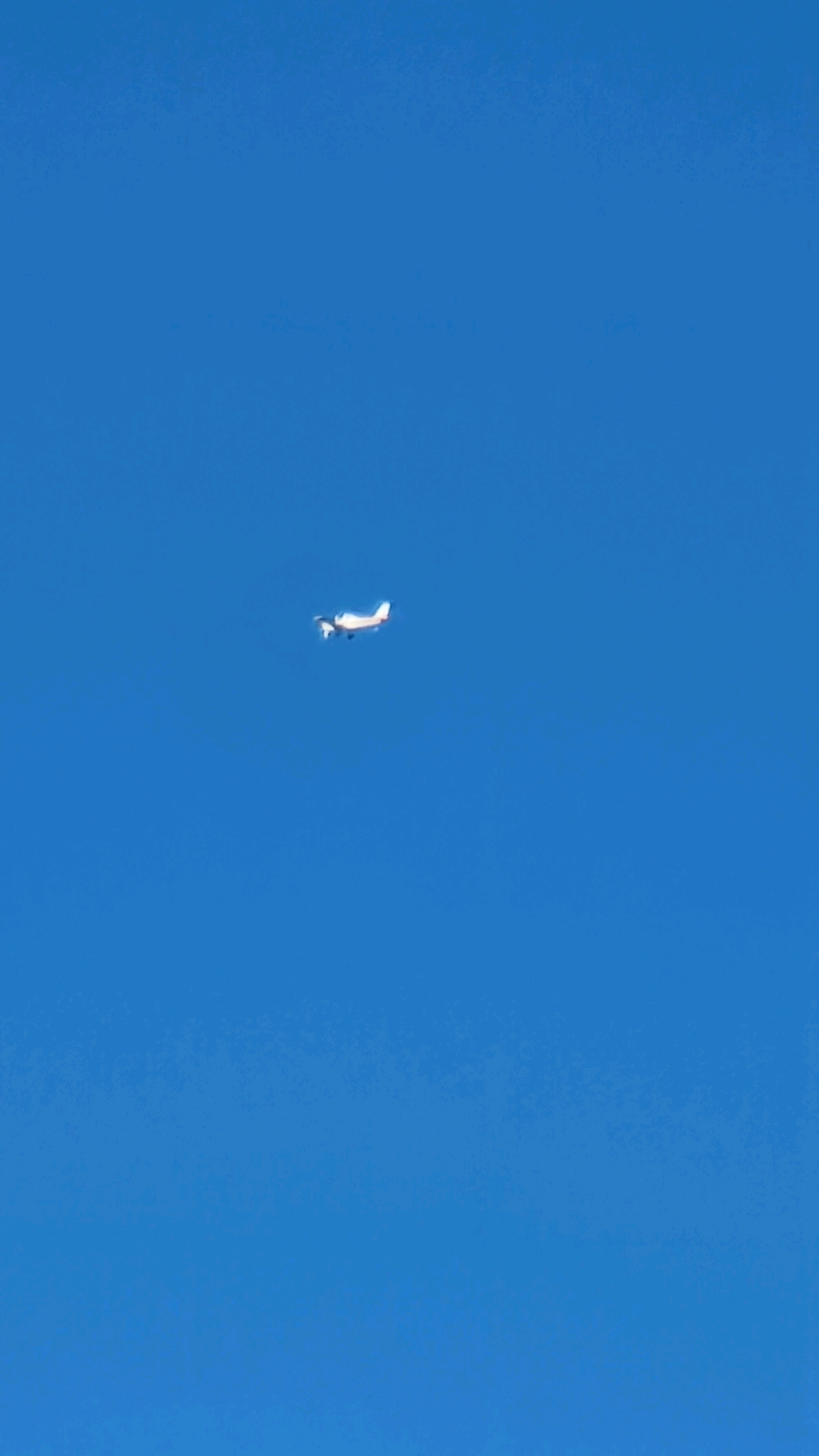 A single-engine airplane with white and red accents is captured mid-flight against a clear, deep blue sky. The aircraft is positioned slightly right of the vertical center and to the left of the horizontal center of the frame. Its wings are extended, and the propeller is visible. The airplane appears to be moving away from the viewer. The background is a uniform expanse of blue, indicating an outdoor scene with no discernible landmarks or structures from Sant Joan Les Fonts, Spain, visible in the image.