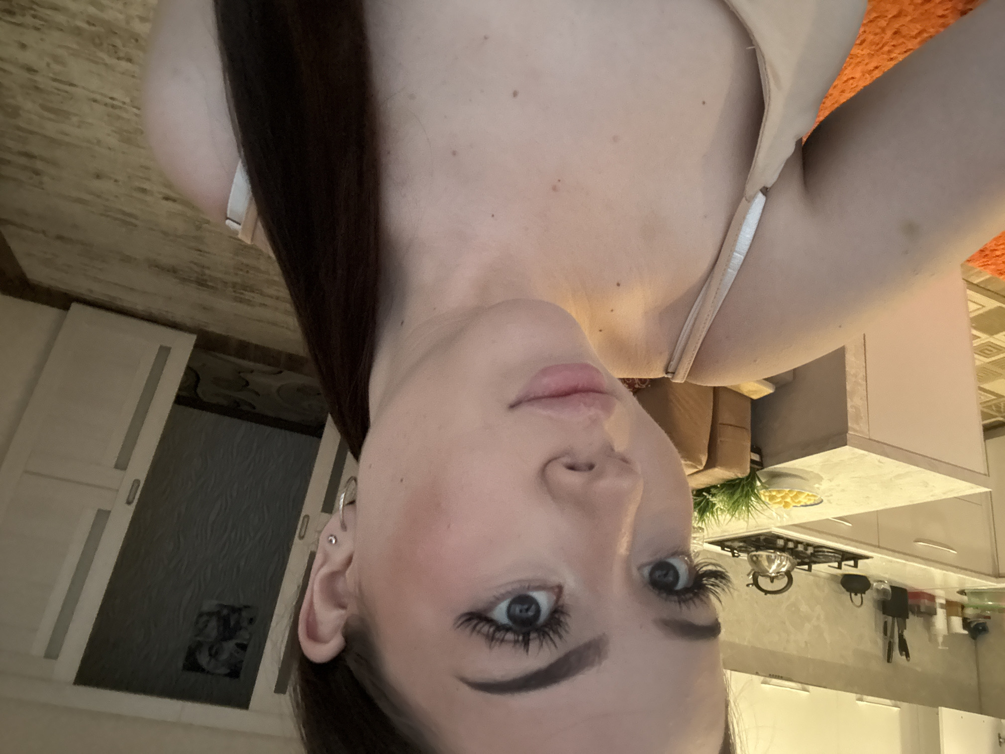 The media file is an inverted, close-up indoor selfie featuring a young woman. The woman has long, dark hair and fair skin with several small moles or freckles on her chest and neck. She is wearing a light-colored (possibly beige or nude) bra or top with thin straps. Her makeup includes prominent long eyelashes and dark, defined eyebrows, with a neutral expression and slightly parted lips. Her left ear has multiple piercings, including hoops and studs.
The background setting appears to be a residential kitchen or an open-plan living area that includes a kitchen. Visible kitchen elements include a gas stovetop with a kettle, a bowl containing yellow spherical items (possibly fruit or snacks), hanging kitchen utensils, and various jars or containers on countertops. There are modern white or light-colored kitchen cabinets. To the left, a portion of a light-colored sofa or chair is visible. Further in the background, a dark door or panel with a patterned surface and a picture/poster is seen. The flooring is a wooden or wood-patterned surface. The overall lighting suggests an indoor setting, likely during daytime or under artificial illumination. No discernible text is visible. The location context provided for this image is Naberezhnye Chelny, Russia.