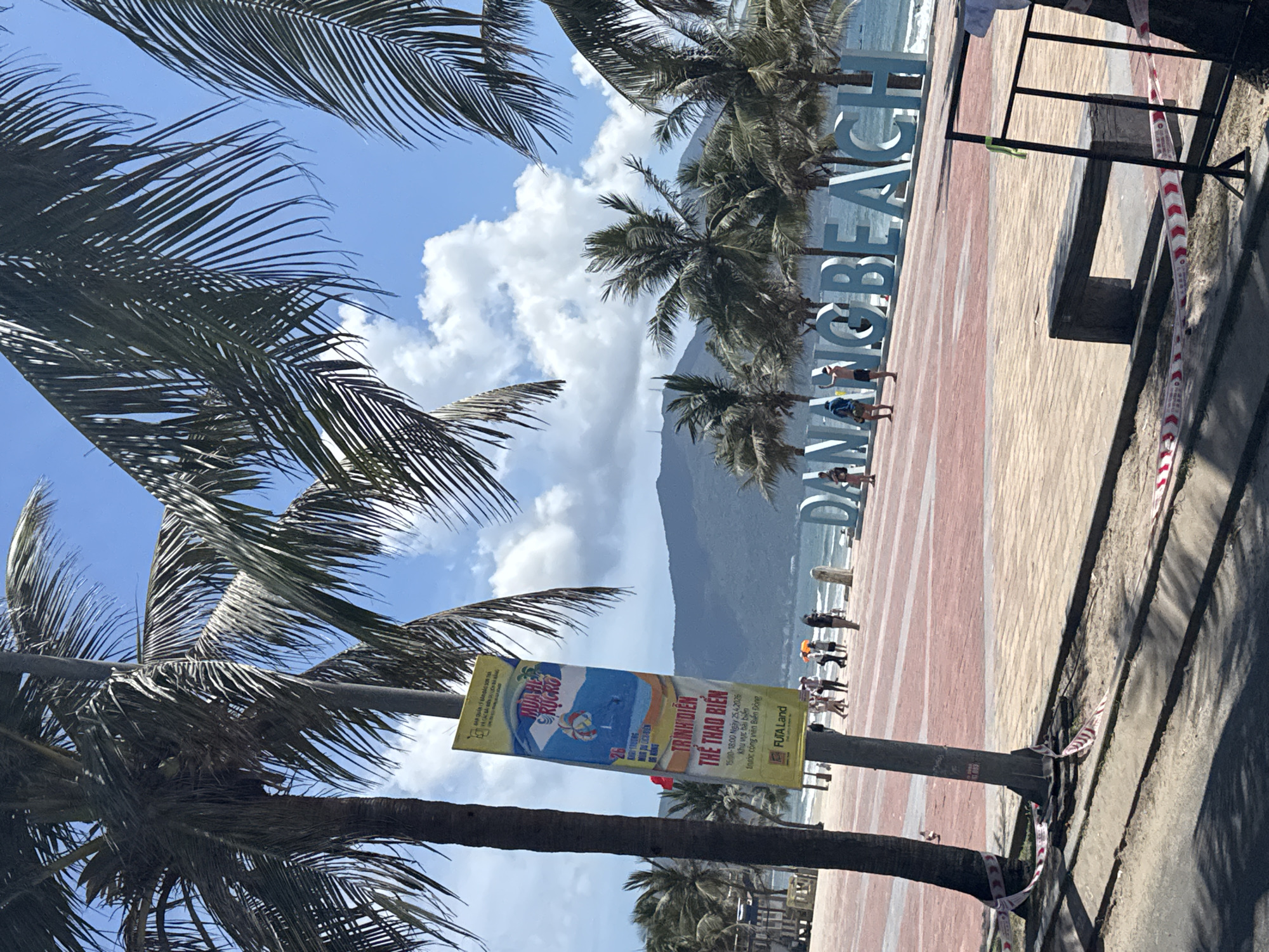 This is a photograph taken at Da Nang Beach in Da Nang, Vietnam. The image is oriented sideways. The beach has a sign that says "DANANG BEACH". There are palm trees along the beach. There are some people walking on the beach. There is a poster in Vietnamese attached to a tree. A mountain can be seen in the background. There is red and white tape on the side of the beach. The sky is blue with white clouds.
