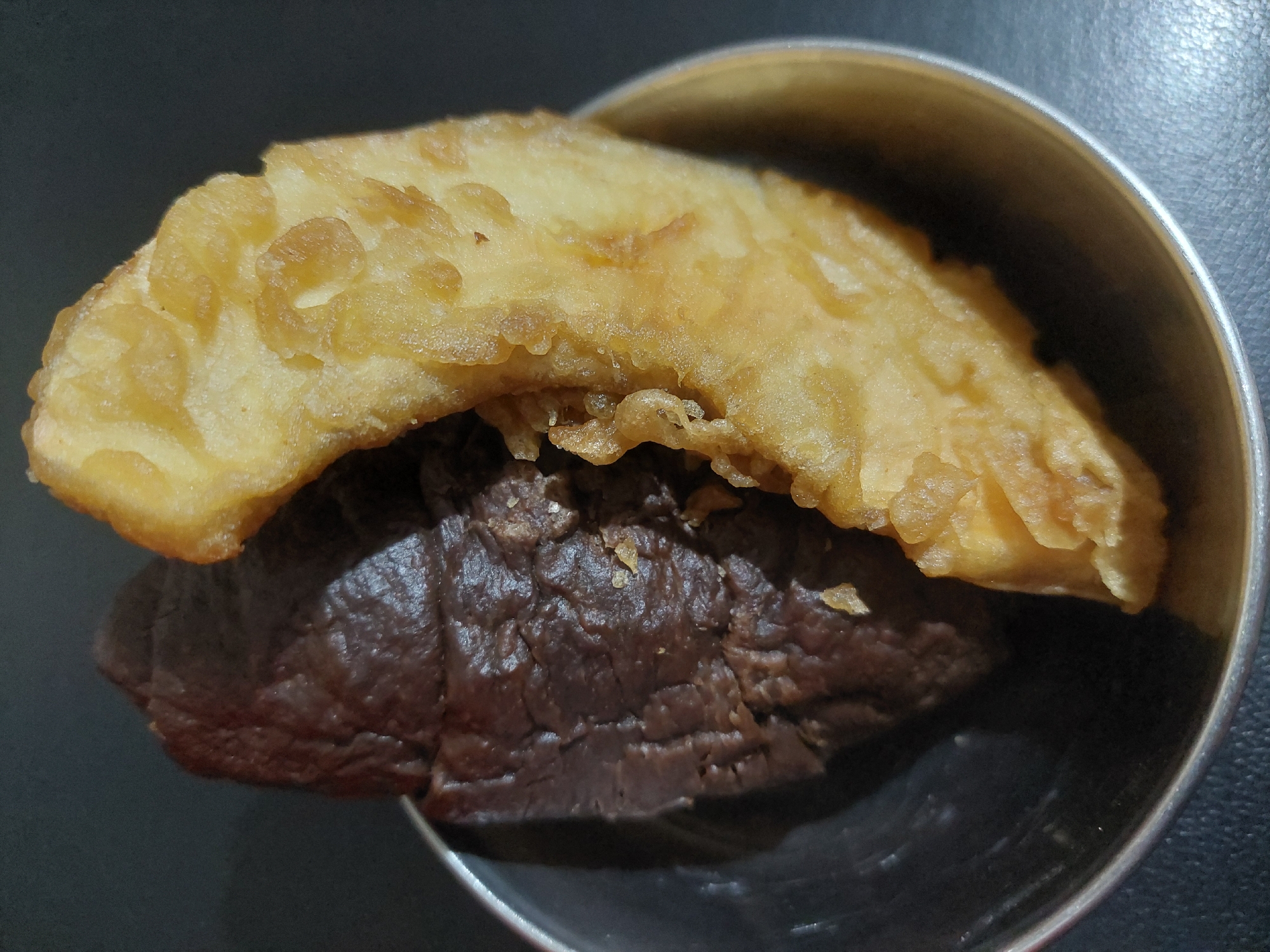 In a small, metallic bowl, two pieces of food are positioned side-by-side against a dark, matte background. The food item on the left is dark brown and has a rough, textured surface, suggesting it might be a piece of dark chocolate or a dense brownie. The food item on the right is golden yellow and appears to be fried, with a crispy, uneven texture, possibly a piece of fried banana or a fritter. The bowl itself is partially visible at the bottom and side of the frame, reflecting some light. The overall scene is a close-up shot focused on the food items. There is no indication of the city of Jelutung, Indonesia, in the image.
