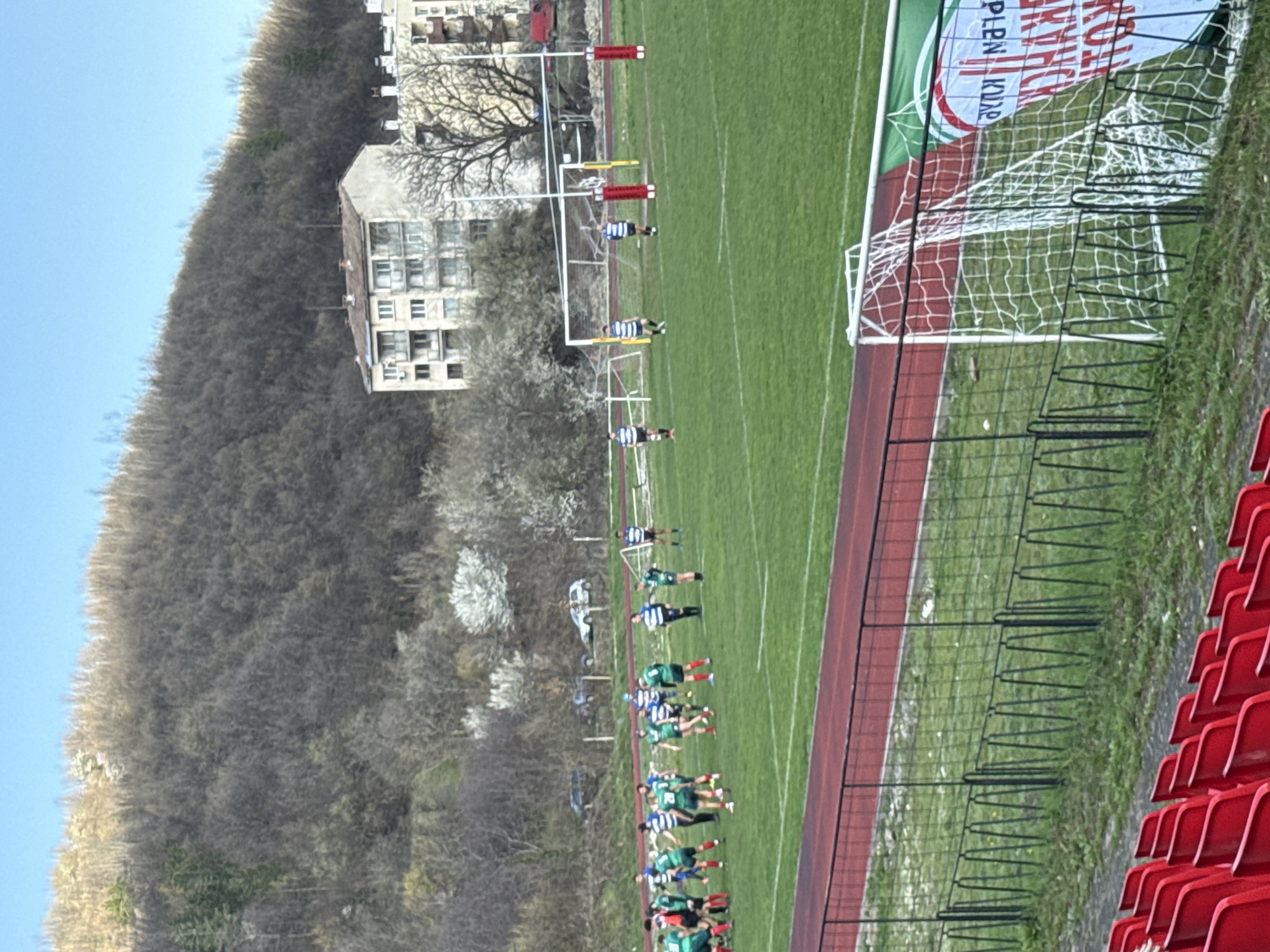 The image depicts a rugby field in Berkovitsa, Bulgaria. On the green pitch, two teams line up facing each other, one in green and red jerseys, the other in blue and white striped jerseys. A white goalpost with a net stands at one end of the field, with a banner bearing the words "KITH CYKOB" written in Cyrillic and "PLEVEN" underneath. A red running track borders the field. Beyond the track, red stadium seats are visible. In the background, a bare forest-covered hillside rises, with a multi-story building visible among the trees. A white American football-style goalpost stands further down the field. The sky is pale blue.