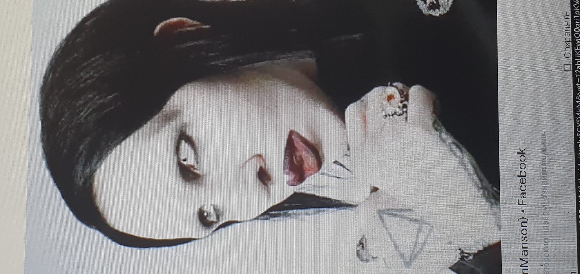 The image shows a close-up, head-and-shoulders portrait of a person with pale skin, dark hair, and striking white eyes with black eyeliner. The person's lips are painted a deep red, and they are slightly parted, with the tongue visible. They are wearing dark clothing and a ring with a large, multi-colored stone on their right hand. The image appears to be a screenshot or a photograph of a screen, as indicated by the grid pattern and text at the bottom, which includes "Manson) Facebook" and a URL. There is no indication of the city of Bat Yam, Israel, in the image or the associated text.