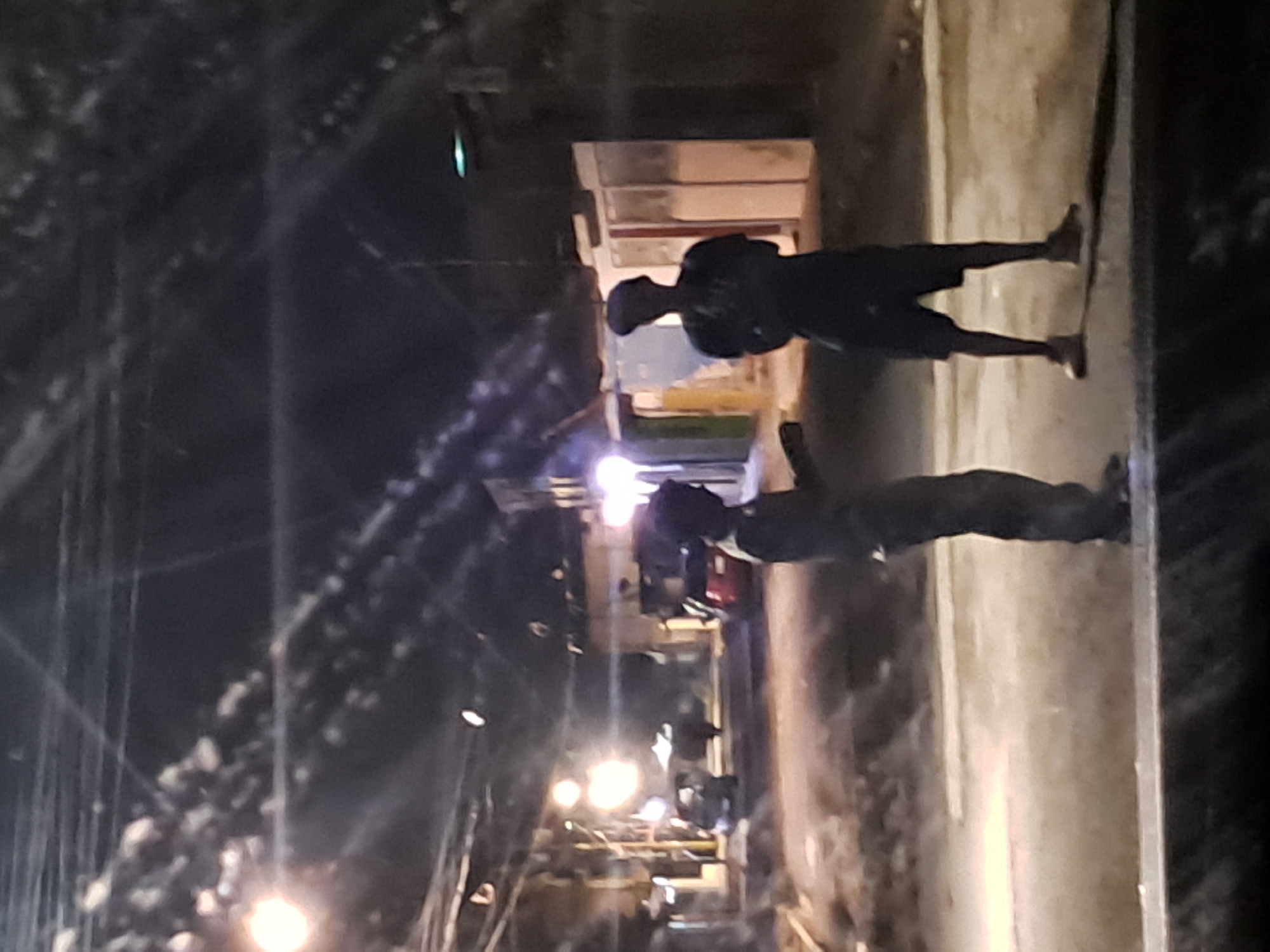 The image depicts a nighttime street scene in Cumana, Venezuela, likely a residential or commercial area given the presence of buildings and street lighting. Two individuals are visible standing in the middle of the street, silhouetted against the ambient light. The person on the left is facing towards the right, appearing to be speaking or gesturing with their hand. The person on the right is facing forward, with their arms crossed. Cars are present in the background, one appearing to be a red sedan. The overall atmosphere is dimly lit, with streetlights casting a glow on the wet or reflective street surface. The scene suggests a casual or perhaps impromptu gathering on the street at night.