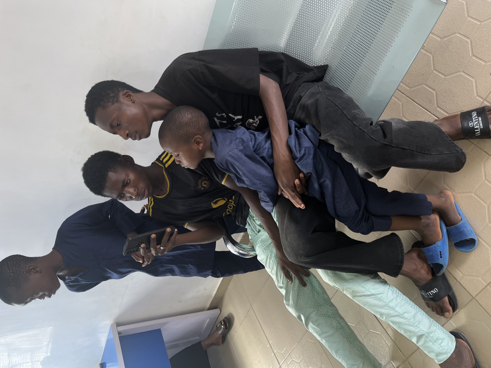 Four Black individuals are seated on a bench. One is holding a phone, two are wearing sandals, and one is wearing blue slides. The walls are white and the flooring is light-colored with a pattern. This image was taken in Gura Top, Nigeria.