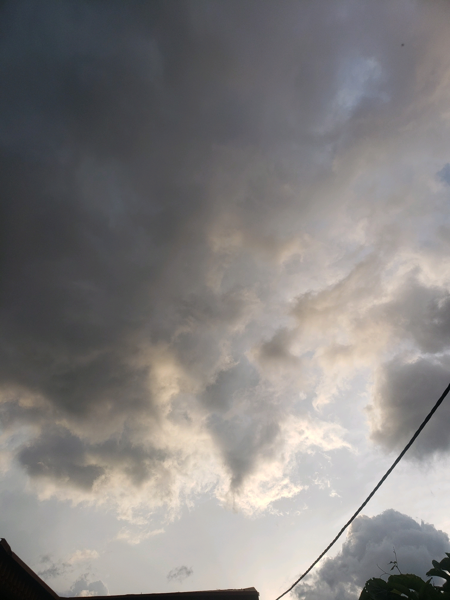 A dusky sky with dramatic, dark grey clouds dominates the scene. To the left, a sliver of a brown rooftop is visible, suggesting an urban setting. A thin, black electrical wire cuts diagonally across the frame from the upper right to the lower right. Patches of lighter, brighter clouds break through the darker mass, hinting at diffused sunlight. The overall atmosphere is somber and possibly foreboding, indicative of impending rain. This scene could be observed in Sabon Tasha, Nigeria, a location characterized by its tropical climate where such cloud formations are common.