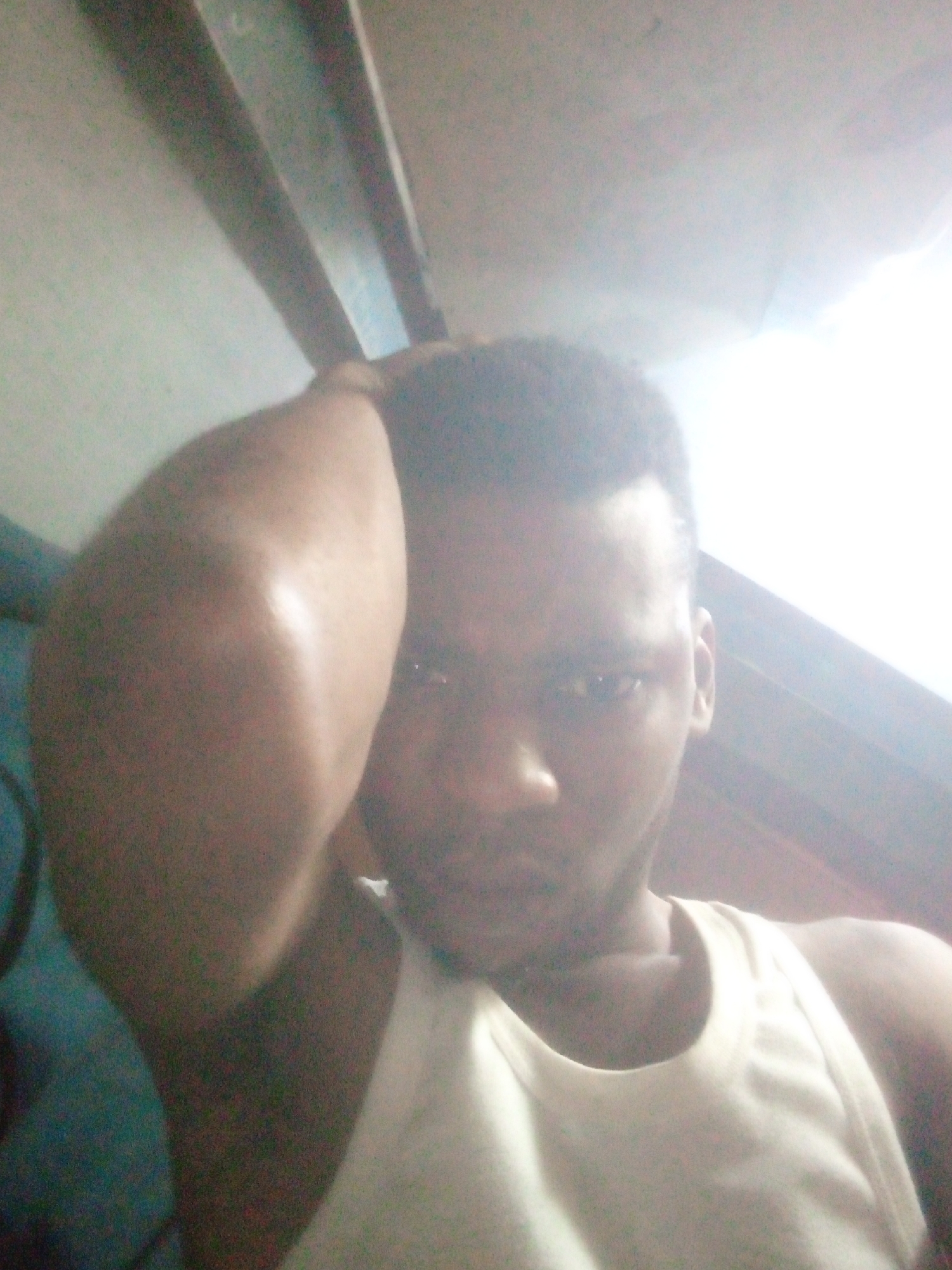 A close-up, poorly lit selfie of a dark-skinned man wearing a white sleeveless shirt. His right arm is raised, with his hand seemingly touching his head. The background includes what appears to be wooden beams or slats, possibly part of a ceiling or structure. The location is unspecified and unknown.