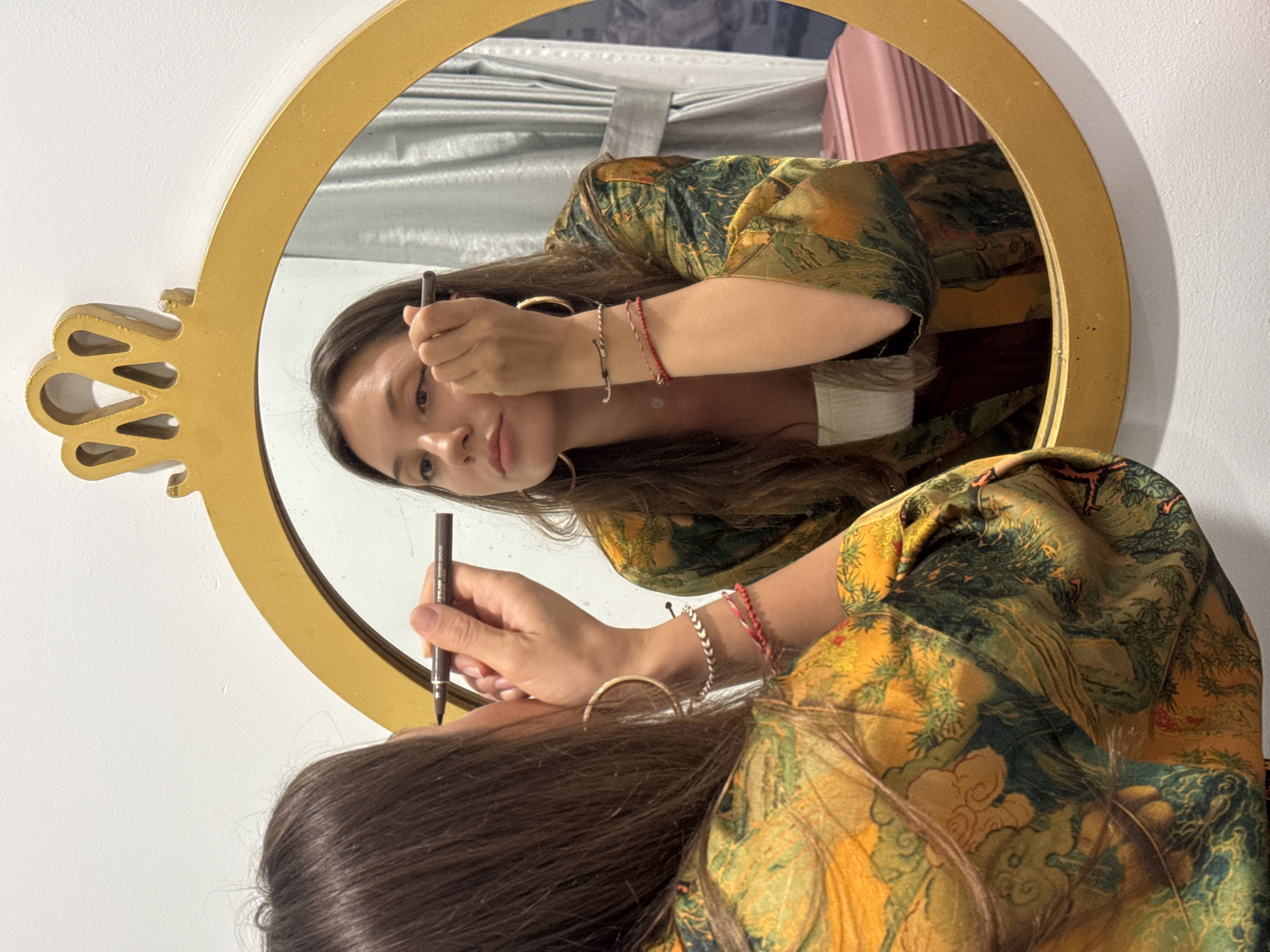 A woman is shown in a mirror, applying eyebrow makeup with a pencil. The mirror has a gold frame and ornate decorative elements. The woman has long brown hair, gold hoop earrings, and wears a patterned green and gold top. The background shows a draped silver fabric and a pink object. The location cannot be determined from the image.