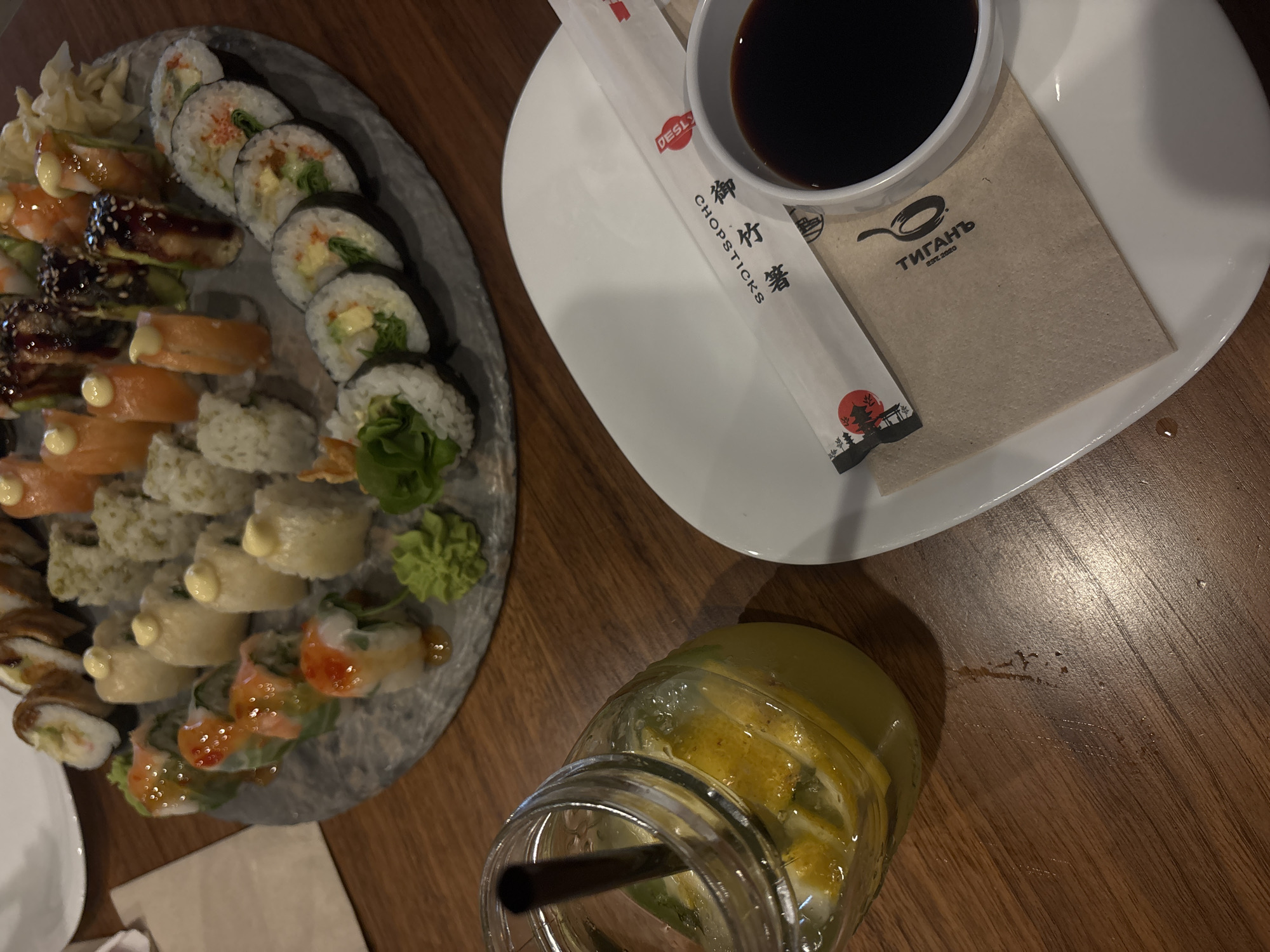 A high-angle shot captures a wooden table setting. A round, grey platter holds a variety of sushi rolls, including maki and nigiri, along with wasabi and ginger. A white plate contains a small bowl of soy sauce, chopsticks in a wrapper, and a tan napkin with black text ("ТИГАНЪ EST. 2020") printed on it. A glass jar filled with a yellow liquid, possibly lemonade or a similar beverage, rests on the table. The location is Varna, Bulgaria, as indicated by the napkin text.