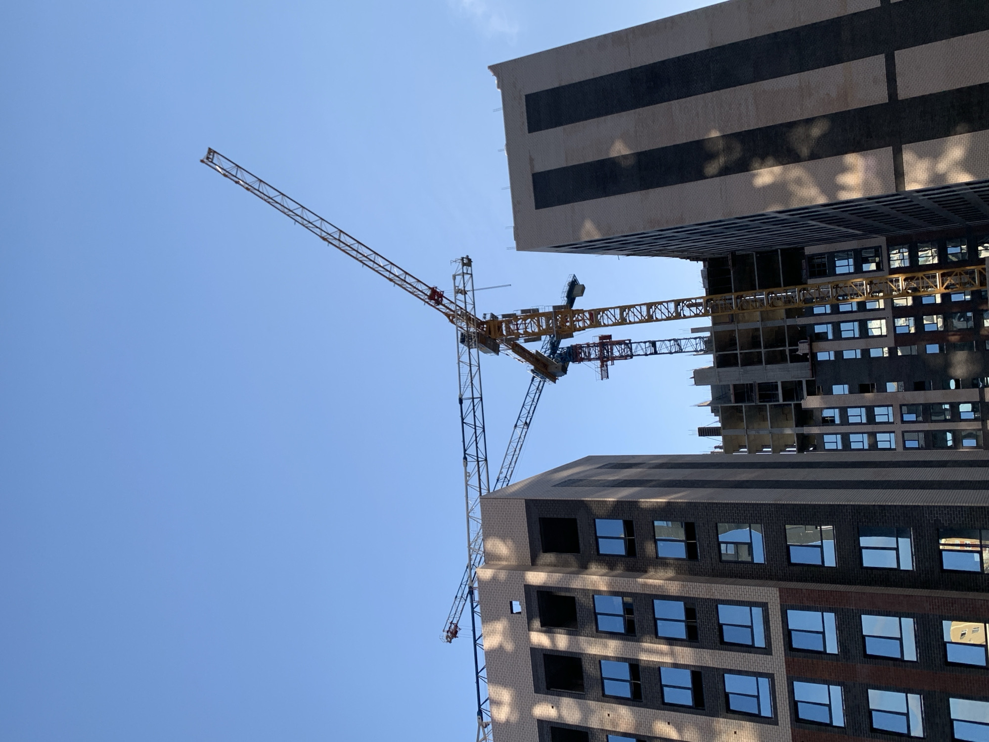 This media file captures a construction scene with two large tower cranes against a bright blue sky. The cranes are in the process of building or extending two modern buildings. The building in the foreground, on the right, is partially constructed and features a dark brick facade with rows of reflective windows. The building on the left appears to be further along in its construction, with a similar facade and visible structural elements. The scene is shot from a low angle, looking upwards, which emphasizes the scale of the buildings and the cranes. The sunlight casts distinct shadows on the facade of the foreground building, suggesting it is midday. The weather appears clear and sunny, typical of a construction day. There are no visible people or text in the image. The location context is Stavropol, Russia.