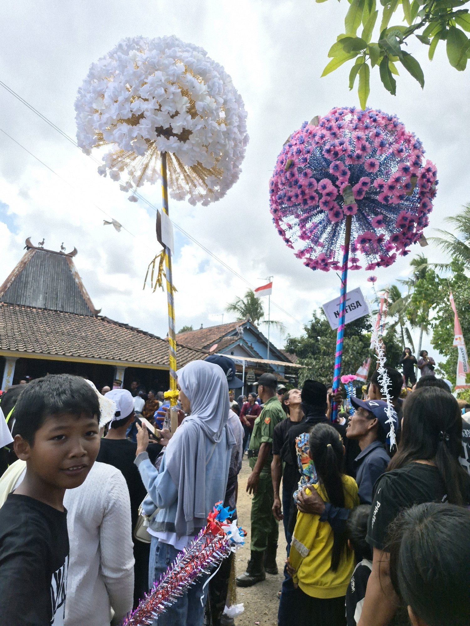 In Grantung, Indonesia, a crowd gathers outdoors under a cloudy sky. Two large, spherical decorations, one white and one pink and blue, are held aloft on poles. The white decoration is made of numerous small white flower-like objects, while the pink and blue decoration features small pink flowers with blue accents. A sign with the text "NFISA" is attached to the pink and blue decoration's pole. In the foreground, a young boy with dark hair looks towards the camera, smiling. He is wearing a black t-shirt. Behind him, a person in a white sweater is visible. Further back, a person in a light blue headscarf and a grey outer garment is holding the pink and blue decoration. Other people in the crowd are dressed in casual clothing, with some wearing hats and head coverings. Traditional Indonesian buildings with tiled roofs are visible in the background, along with palm trees and foliage. A red and white Indonesian flag is also present. The overall scene appears to be a festive or ceremonial gathering.