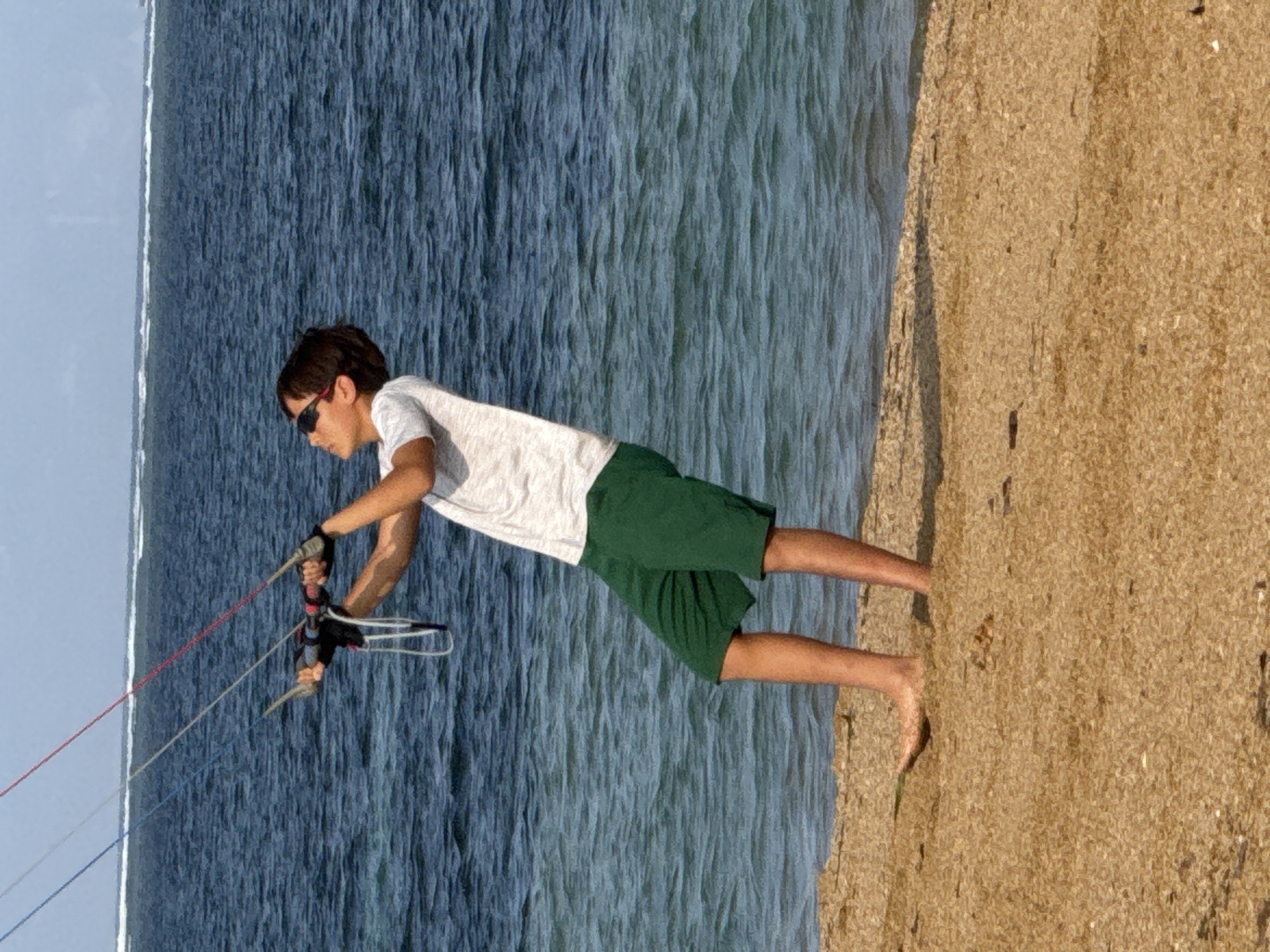 A young male, wearing a white t-shirt, green shorts, and sunglasses, stands on a sandy beach, holding onto the control bar of a kiteboarding setup. The kite lines extend upwards, out of frame. The sea, with moderate waves and a distant horizon, occupies the background. The sand is light brown and appears somewhat textured. The image is captured from a mid-range perspective, focusing on the individual and the immediate shoreline. The scene is set outdoors, likely during daylight hours, under a clear or partly cloudy sky. The location is identified as Denpasar, Indonesia, implying a tropical coastal environment.