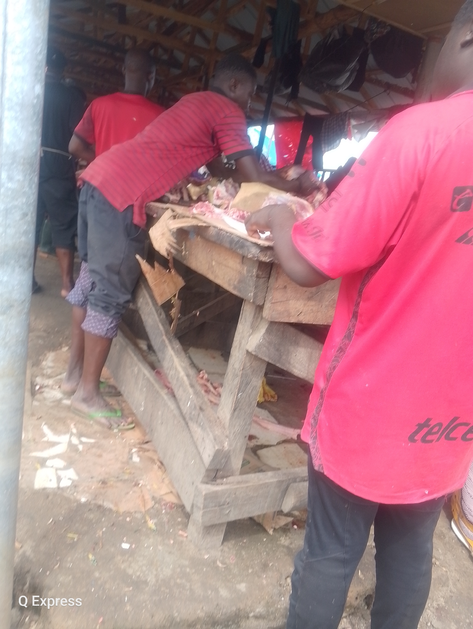 The image captures a scene in a bustling outdoor market in Sabojibi, Nigeria, with a focus on butchery activities. In the foreground, a person wearing a vibrant red t-shirt with the "telcel" logo on the back is visible from the waist down, standing with their back to the camera. They are dressed in dark trousers. To their left, another individual in a red and black striped t-shirt and dark shorts is actively engaged in butchery work. They are hunched over a wooden table laden with meat. Behind them, a person in a red t-shirt and dark shorts is also present. The environment appears to be a rustic, open-air market with a wooden structure serving as a stall, covered by a corrugated metal roof. The ground is dirt, with scattered debris and pieces of meat indicating ongoing commercial activity. The overall impression is one of a functional and active marketplace.