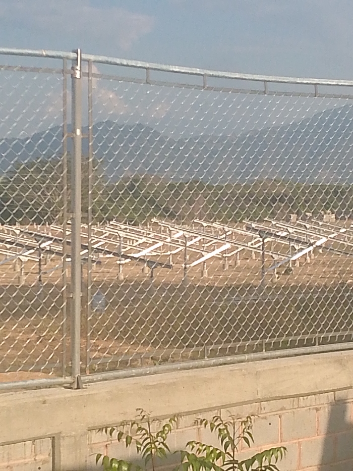 A chain-link fence dominates the foreground, obscuring a wide, arid landscape. Rows of metal structures, presumably the support frames for solar panels, extend across the flat expanse. A line of trees separates the plains from distant, hazy mountains under a pale blue sky. A concrete wall, partially visible, anchors the fence. Small plants grow near the wall.