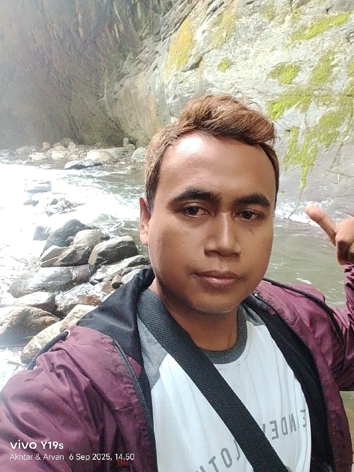 A selfie-style portrait of an adult male is presented, taken outdoors in a natural environment identified as being in the city of Sukadana, Indonesia. The individual, appearing to be of Southeast Asian descent, occupies the foreground, looking directly into the camera with a neutral expression. He has light brown hair, styled with volume on top, and is wearing a white t-shirt with grey raglan sleeves, overlaid by a dark maroon hooded jacket with a visible black inner lining. A black strap, likely from a bag, crosses his chest. His right hand is raised to shoulder height on the right side of the frame, with the index finger pointed upwards and slightly bent, and the thumb extended.
The background features a flowing body of water, possibly a stream or river, with numerous smooth, greyish-brown rocks scattered within and along its banks. Behind the water and rocks, steep, moist rock faces or cliffs are visible, exhibiting dark, wet sections and patches of light green moss or vegetation. The lighting suggests daylight conditions. The image metadata indicates it was captured with a "vivo Y19s" device on "6 Sep 2025" at "14.50" and includes the text "Akhtar & Arvan".