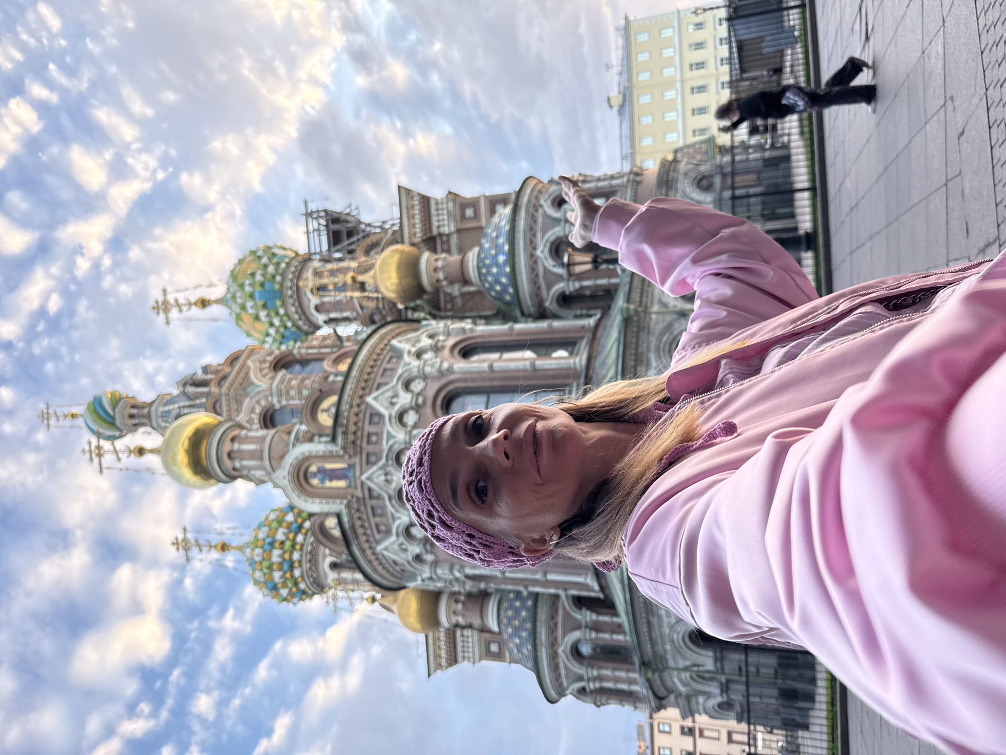The image shows a woman in the foreground, taking a selfie with a historic church in the background. The church, the Church of the Savior on Spilled Blood, is a prominent landmark in Saint Petersburg, Russia, known for its intricate architecture and colorful onion domes. The woman is wearing a pink jacket and a matching headscarf, and she is smiling at the camera. Her right arm is extended, with her hand open, as if gesturing towards the church. In the background, other buildings and a person walking can be seen. The sky is cloudy, with patches of sunlight breaking through. The overall scene depicts a tourist capturing a moment of admiration at a famous historical site.