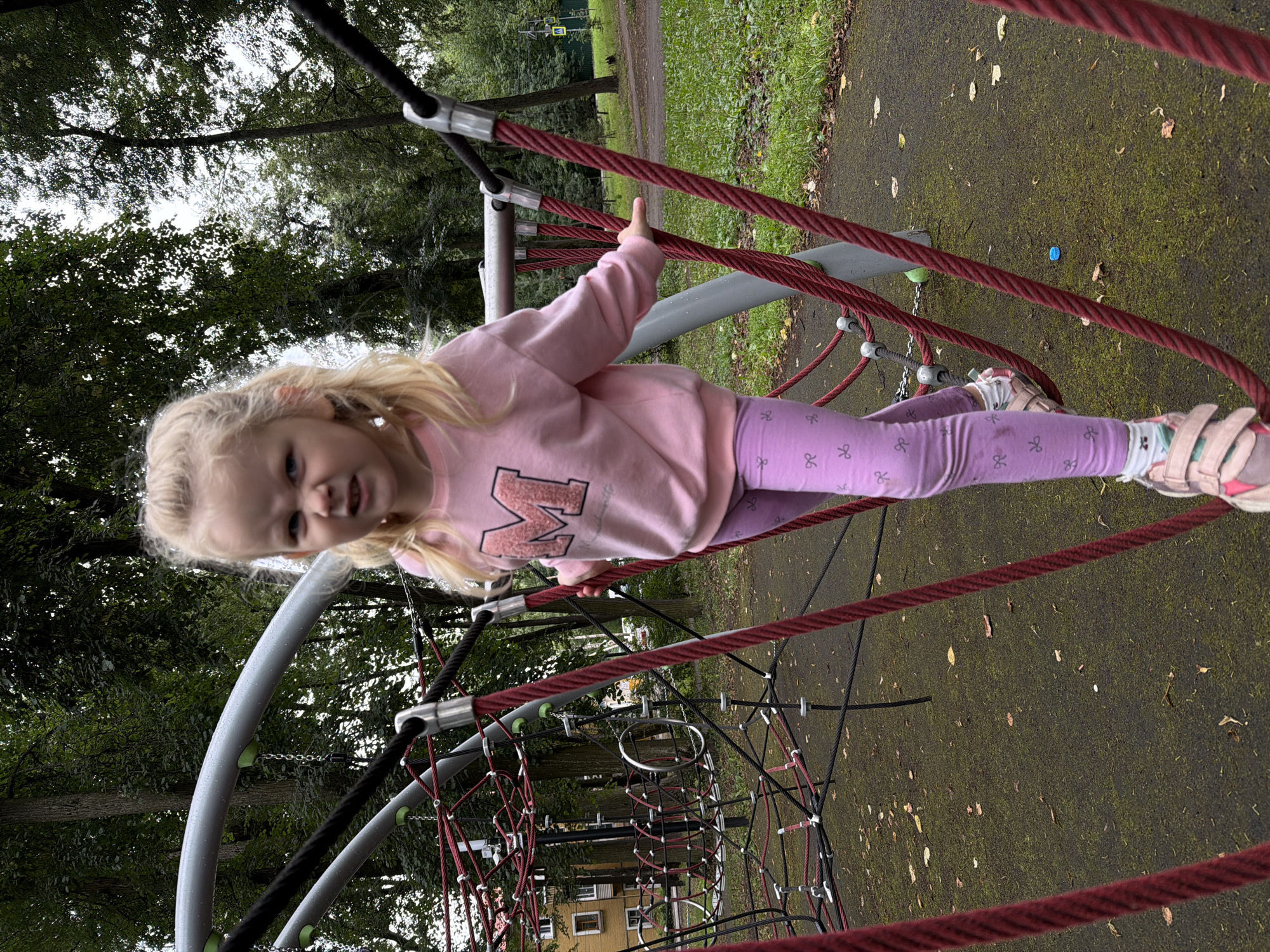 A young Caucasian girl with blonde hair pulled into a ponytail is standing on a rope climbing structure in a park. She is wearing a pink long-sleeved shirt with a large "M" graphic and purple leggings with a bow pattern. Her shoes are pink and white sandals. She is looking towards the camera with a scowling expression. The playground structure is made of thick red and black ropes with metal connectors. The ground beneath her is a dark, textured surface covered in fallen leaves and moss. In the background, there are tall trees with green foliage and a building with yellow siding. The setting appears to be a public park in Malakhovka, Russia, given the presence of the building and the general park environment. The lighting suggests it is daytime, possibly overcast.