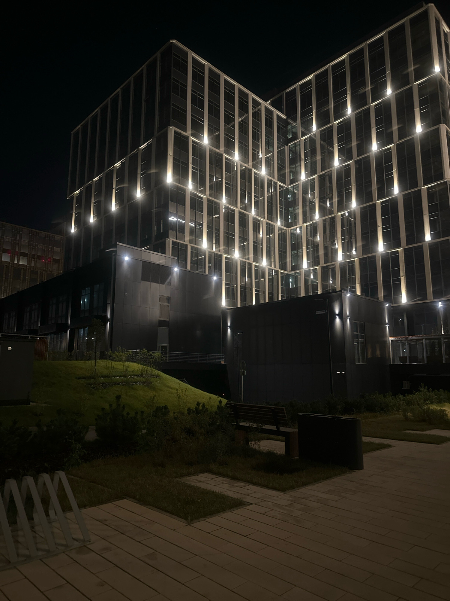 An outdoor night scene in the city of Kommunarka, Russia, features a modern multi-story building with a facade of glass and metal, illuminated by vertical lights. In front of the building is a dark, low-rise structure. A sloped grassy area with bushes and small trees is situated between the building and a paved walkway in the foreground. The walkway has a bicycle rack on its left side. A dark wooden bench and a large dark waste receptacle are placed on the grassy area, close to the walkway. The sky is dark and clear.