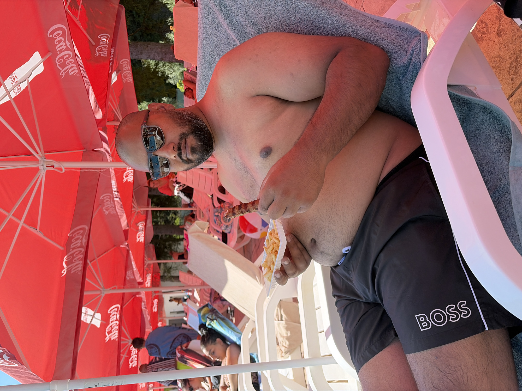 A man is sitting in a lounge chair, wearing black swim trunks with "BOSS" logo on the left leg and sunglasses. He is holding a small white tray with french fries in his left hand and a piece of food in his right hand. He appears to be at an outdoor establishment with red umbrellas in the background, possibly a resort or restaurant in Chiflik, Bulgaria. The weather appears to be sunny.
