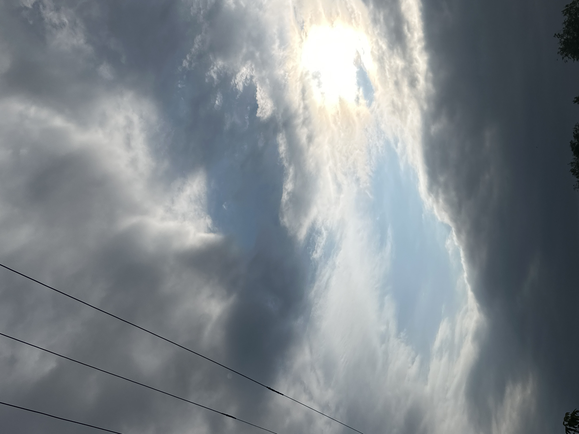 The image displays a dramatic sky filled with clouds. The sun is visible through a break in the clouds, casting a bright, ethereal glow. The clouds themselves vary in color and texture, ranging from dark, ominous storm clouds on the right to lighter, wispy formations on the left. A clear patch of blue sky is visible between the cloud masses. In the foreground, several power lines stretch horizontally across the frame, indicating an outdoor, possibly semi-urban or rural setting. The presence of trees on the far right suggests a natural element within the scene. The lighting and cloud formations create a sense of movement and dynamism in the sky, hinting at changing weather conditions. The overall mood is somewhat dramatic and awe-inspiring, with the interplay of light and shadow on the clouds being a prominent feature. No people are visible in the image, and there is no discernible text. The time of day is likely daytime, given the bright sunlight, but the cloudy conditions could obscure the precise hour.