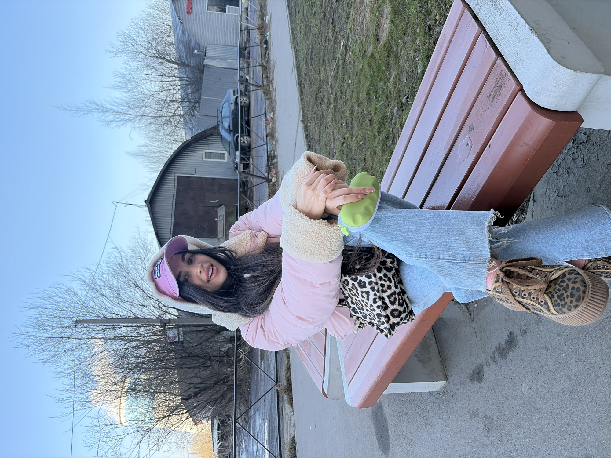 A young female is seated casually on a reddish-brown wooden park bench with white concrete supports. She wears a light pink puffer jacket with a cream fleece-lined hood and cuffs, light blue wide-leg jeans, and leopard print platform sneakers with brown laces. A pink baseball cap with a white logo (identifiable as "Patagonia") covers part of her long, dark hair, which frames a smiling face. Her hands are clasped in front, holding a light green zippered pouch.
The bench is positioned alongside a paved pathway, with a patch of sparse green grass beyond it. In the background, under a clear blue sky, is an urban landscape characterized by bare deciduous trees and several buildings. To the left, a tall, light-colored cylindrical structure, possibly a water tower or industrial tank, is partially visible. Next to it, a large, arched-roof building with corrugated metal siding and a dark brown door stands. To the right, another corrugated metal building with a gabled roof features a red sign displaying the Cyrillic word "ШИНЫ" (Tires). A black sedan with a roof rack is parked between these structures, behind a low, dark metal fence. The ground beyond the fence contains scattered litter.
The presence of the Cyrillic sign "ШИНЫ" confirms the scene is within a Russian-speaking city. The architectural style of the background buildings, which resemble garages or service centers, along with the cylindrical industrial structure, indicates the location is an industrial or commercial fringe area within Saint Petersburg, Russia. The exact street or district is not discernible from the image.